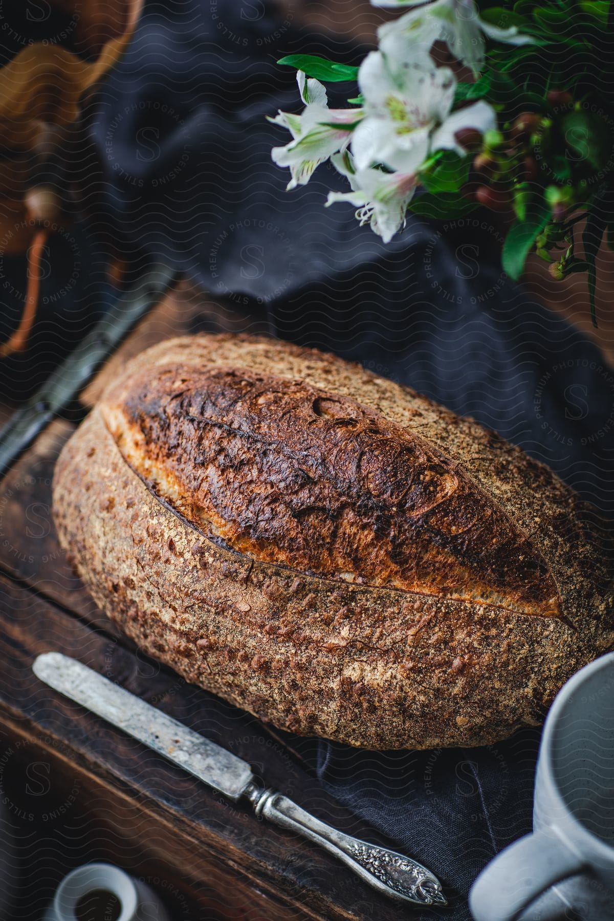 A brown loaf of bread on a wooden surface with a butter knife and flowers nearby