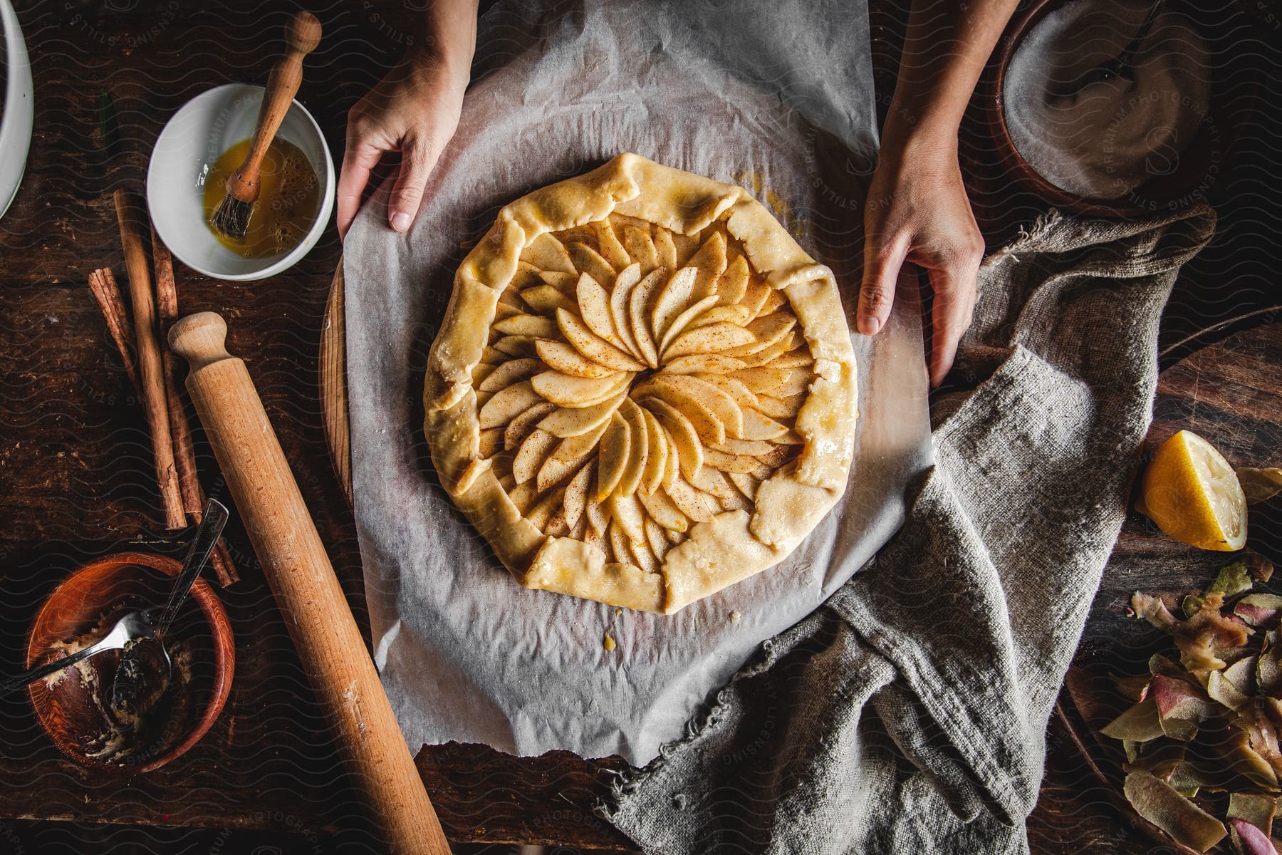 A persons hands displaying a round pie with apple slices on top and kitchen utensils laid on the counter