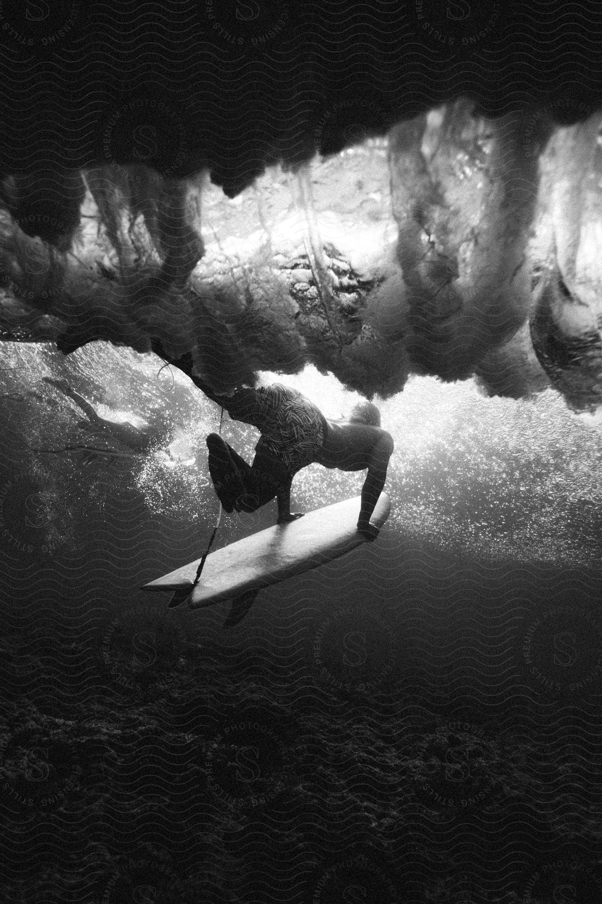 A man swimming under a wave with a surfboard captured in black and white underwater