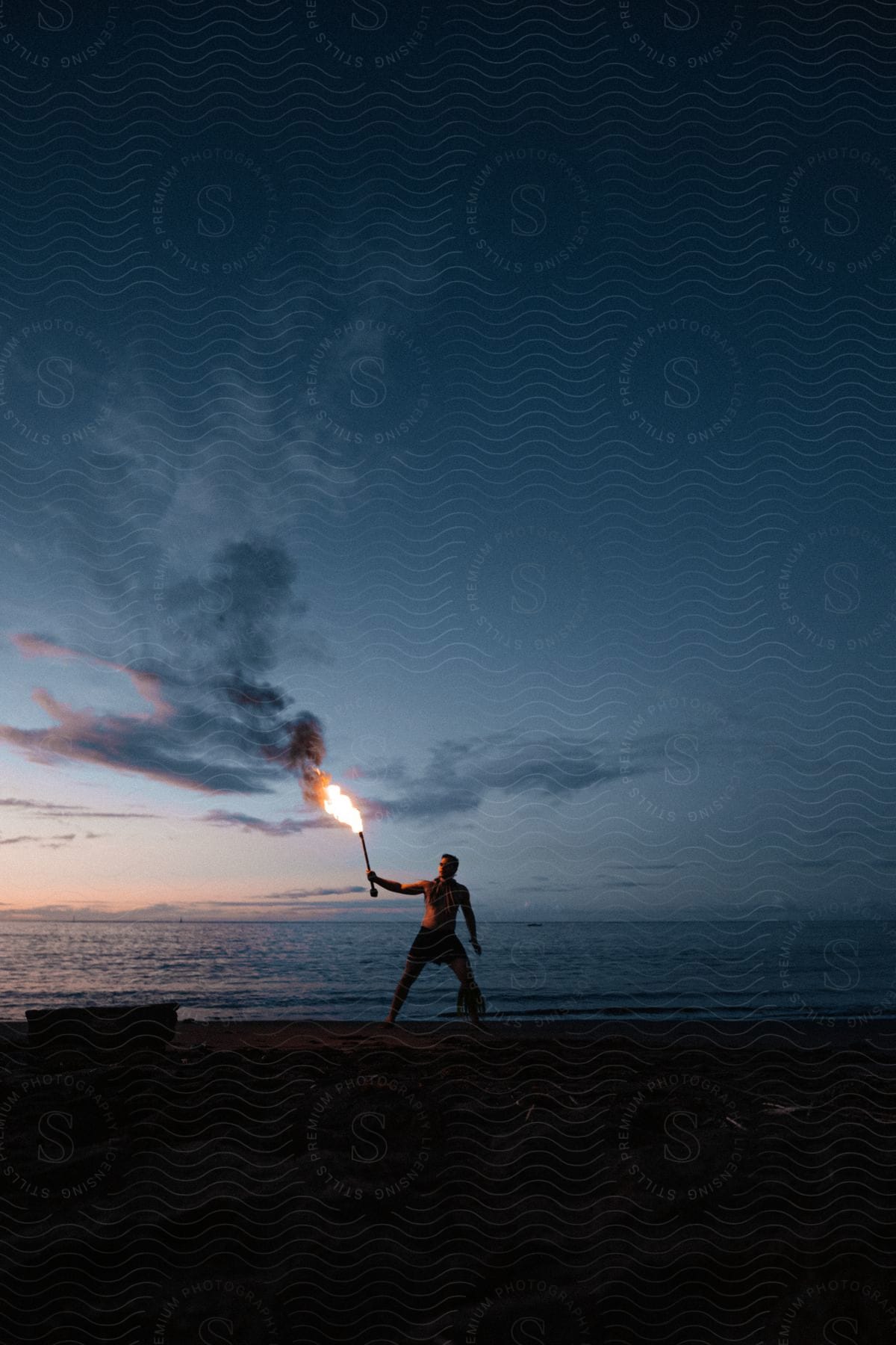 A man stands on the beach holding a flaming torch in the air at dusk or dawn