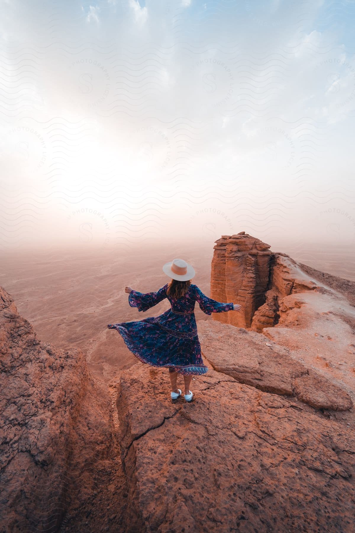 Stock photo of a woman in a white hat and bluepink dress enjoys the breeze from a rocky cliffside on a hazy day