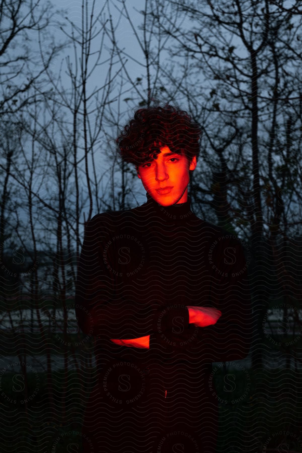 A man in a turtleneck sweater stands with crossed arms in front of leafless trees illuminated by amber light