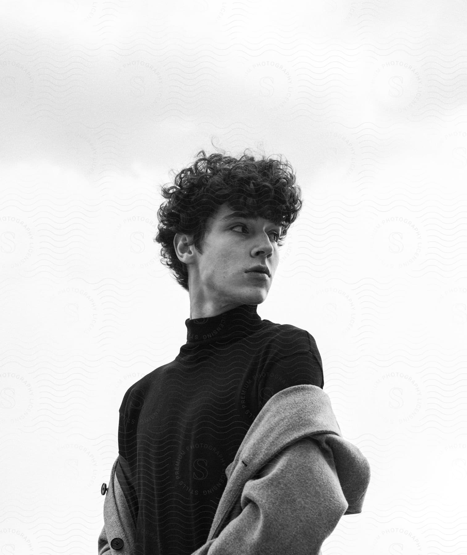 Teenage boy with curly hair wearing a black turtleneck sweater and gray coat looking over his shoulder