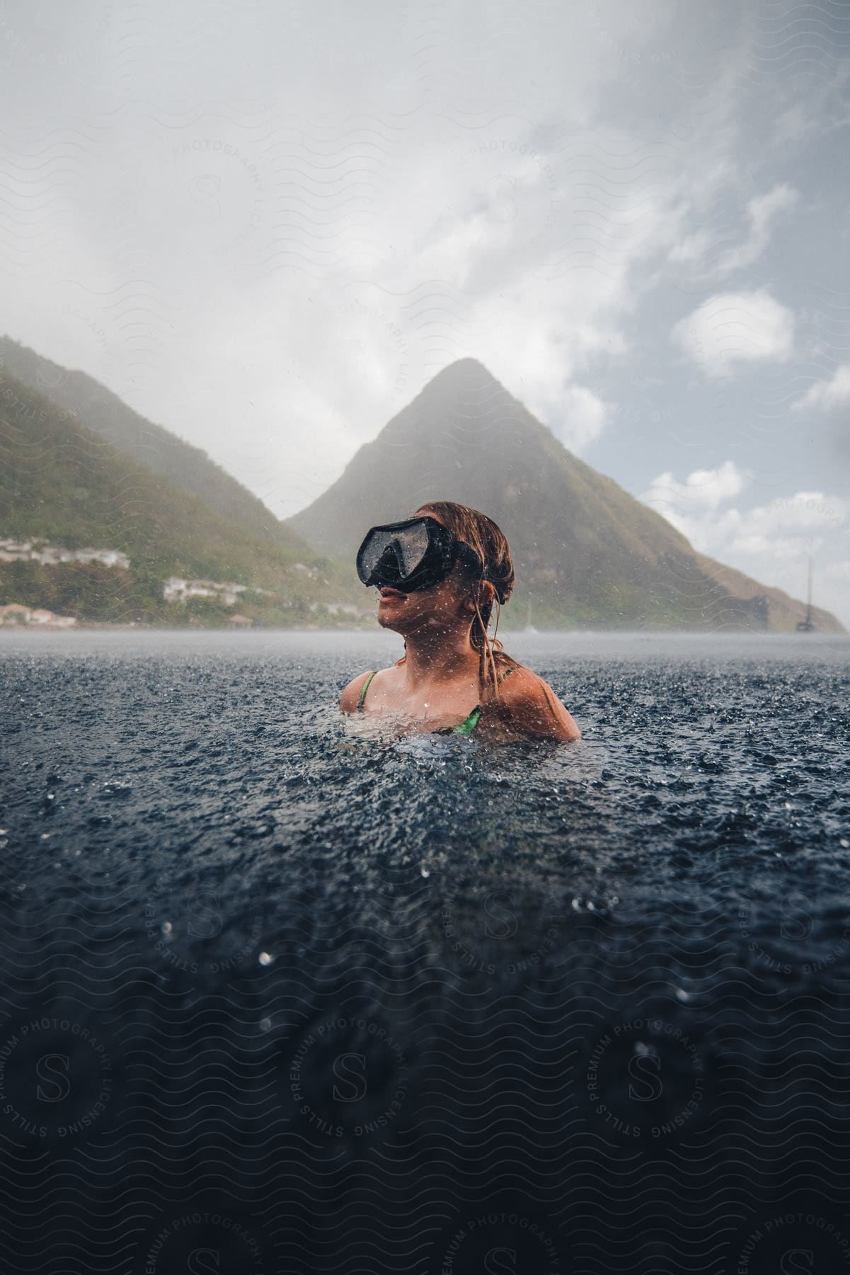 A woman swims in the ocean with goggles on her face raindrops on the waters surface and a mountain in the background as rain falls from cloudy skies