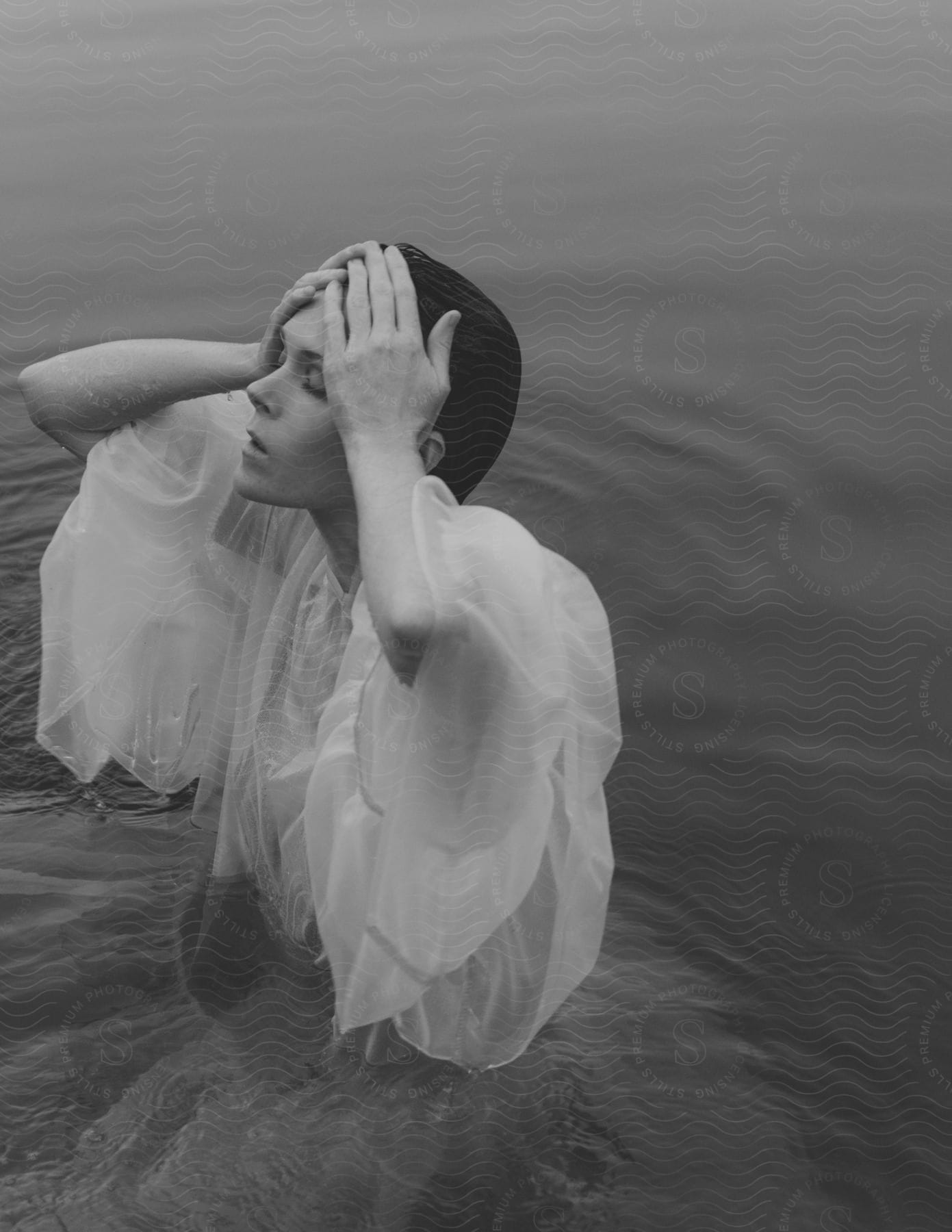 A woman wearing a sheer white dress stands in the water with her hands on her head