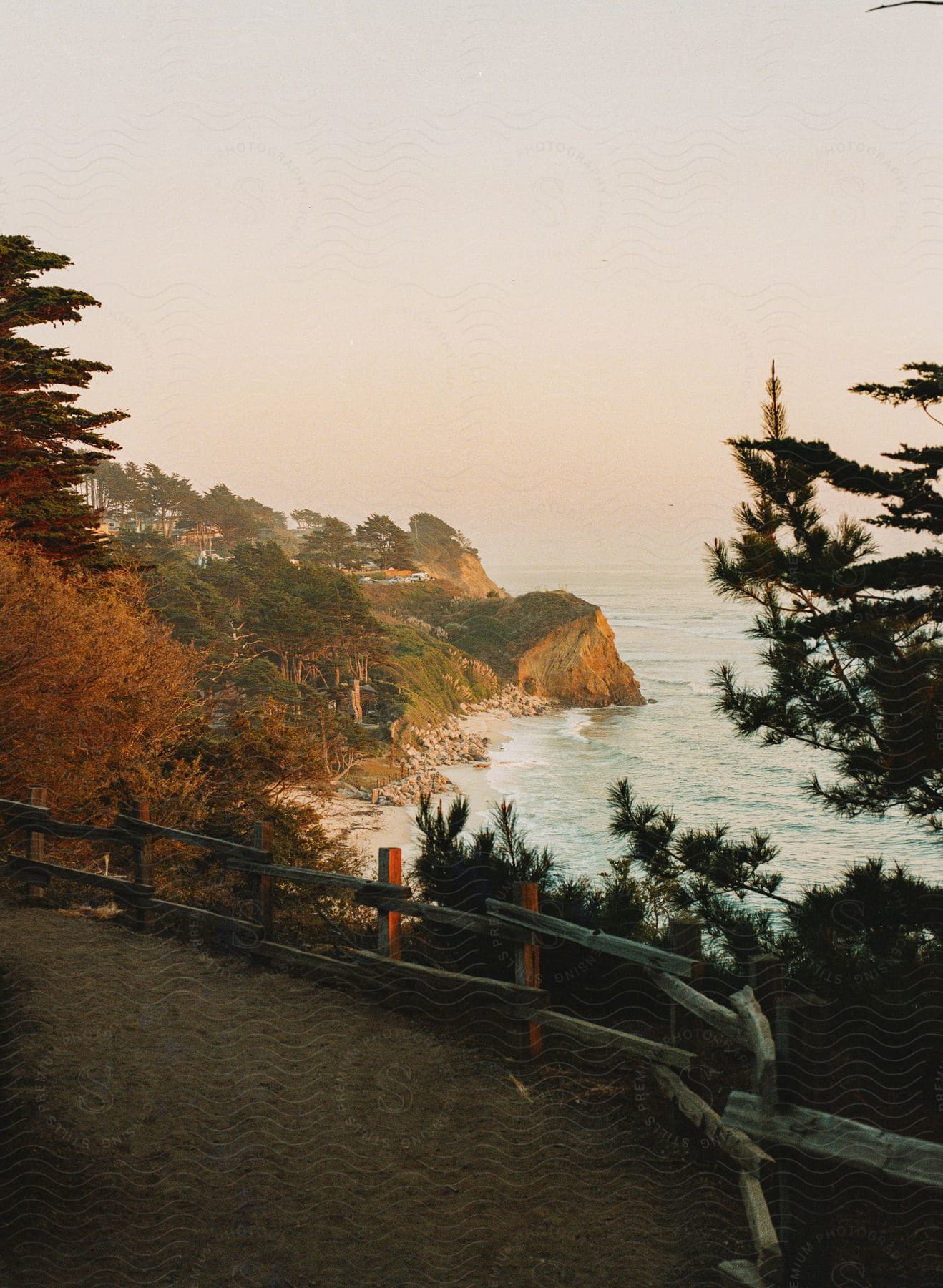 A fence blocks a cliff overlooking the waterfront with trees in fall colors as waves roll into shore