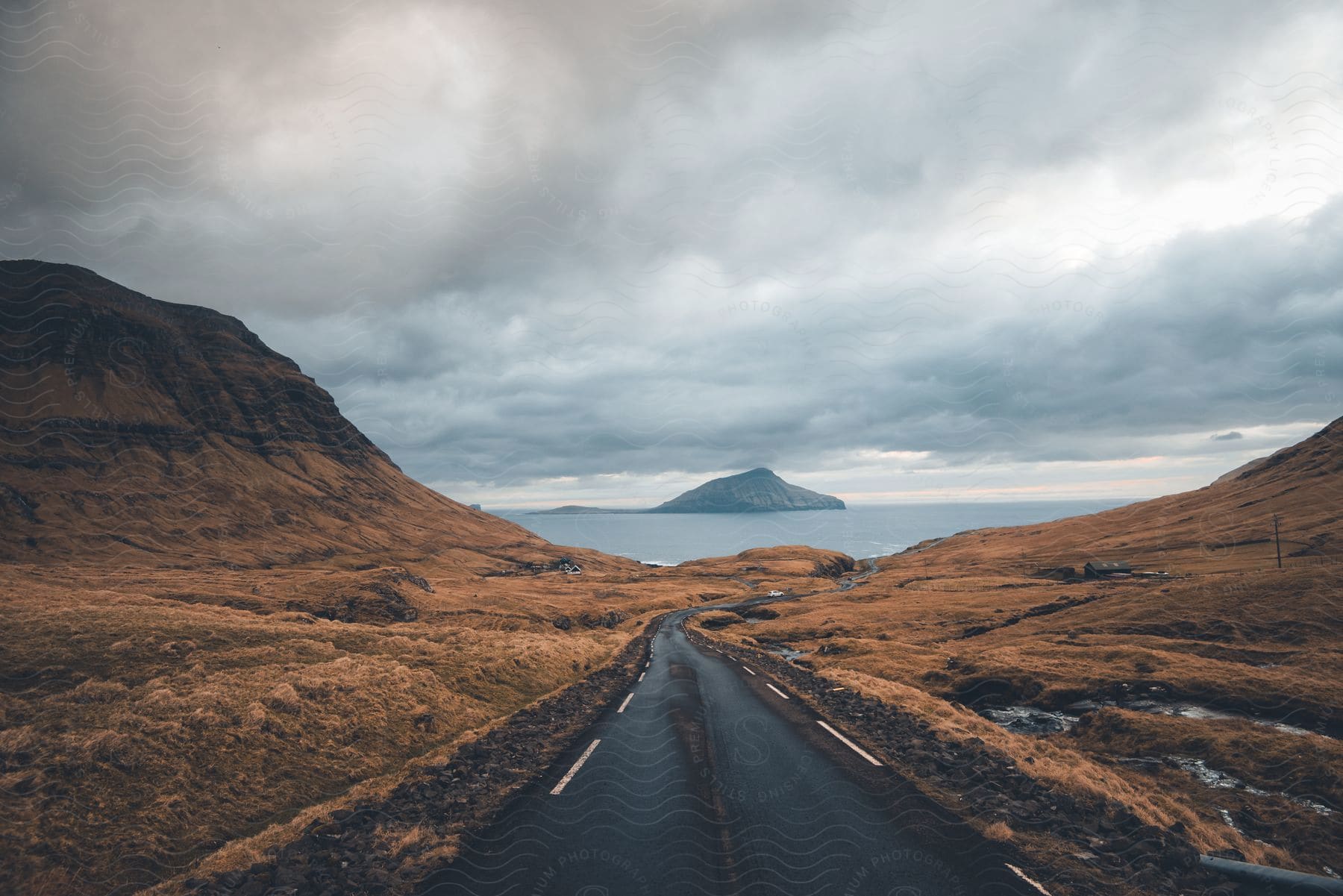 A mountain road leading to the coast with a distant mountain island under a cloudy sky