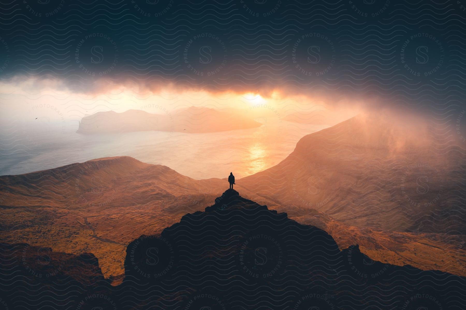 Person standing on a ledge overlooking water and mountains as the sun shines under dark clouds