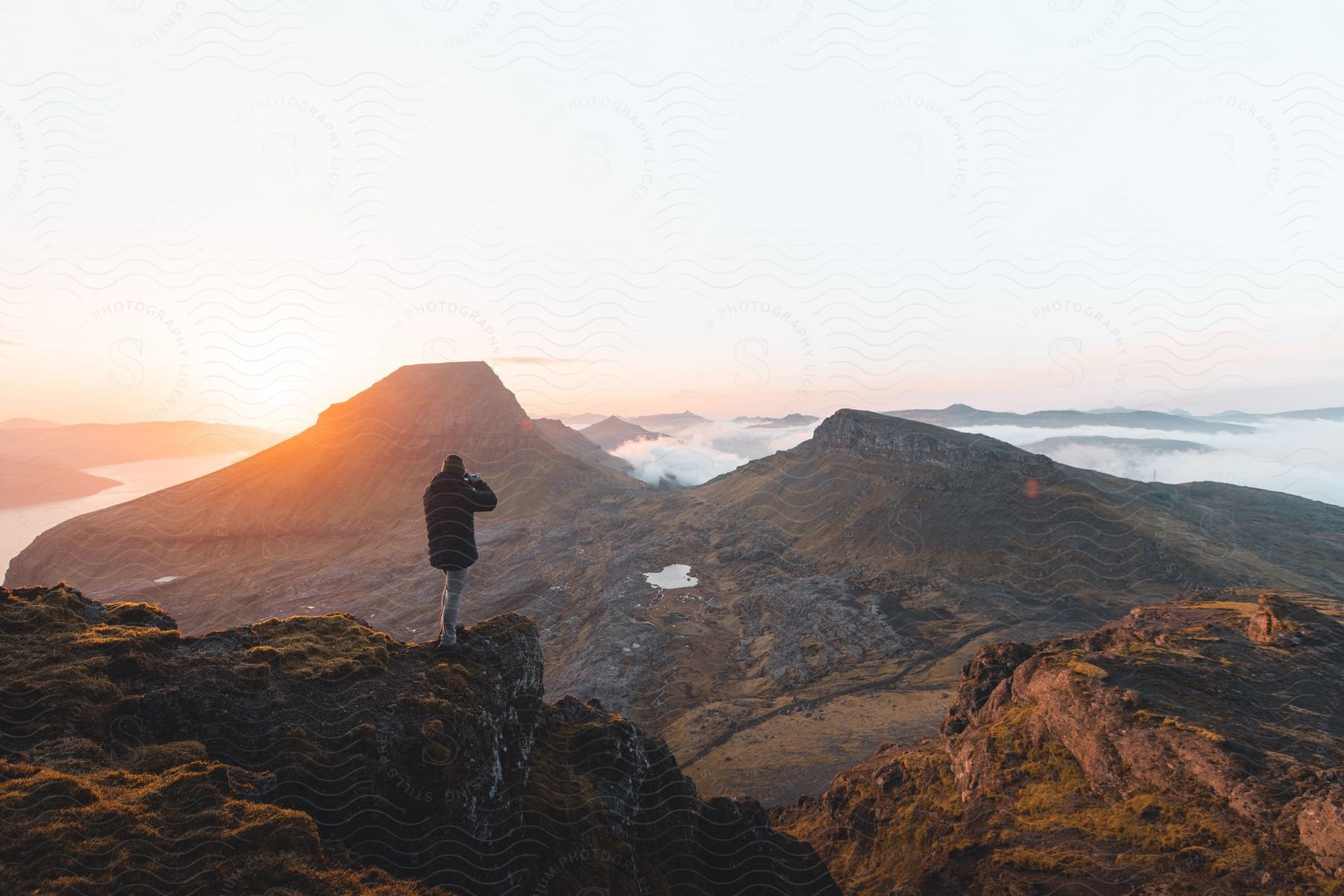 A hiker takes a photo of a mountain valley at sunset with clouds above cliffs and an orange glow on the lake water below