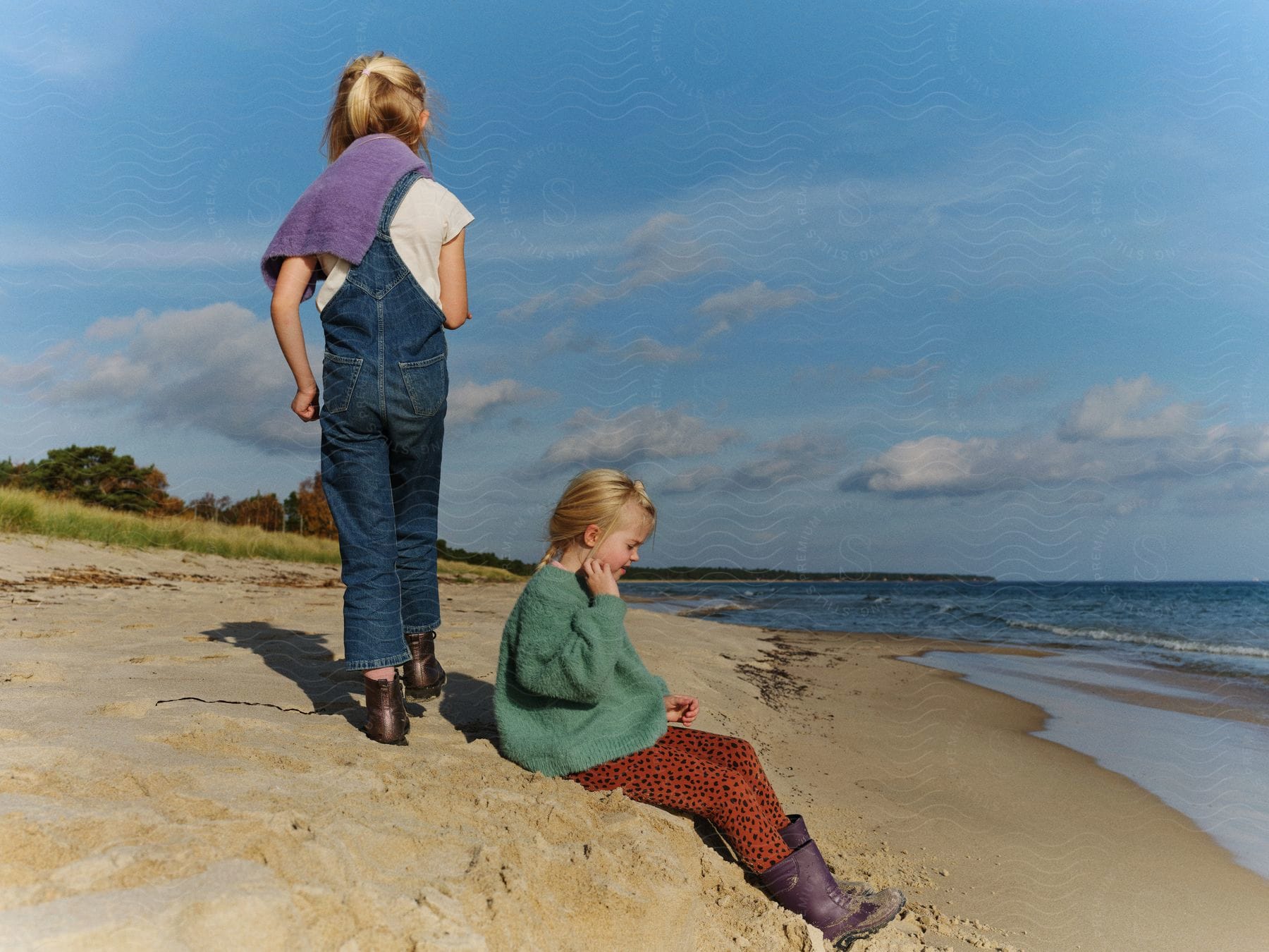 Young girl sitting in sand near water as another girl walks behind her both casting shadows in sand from sun
