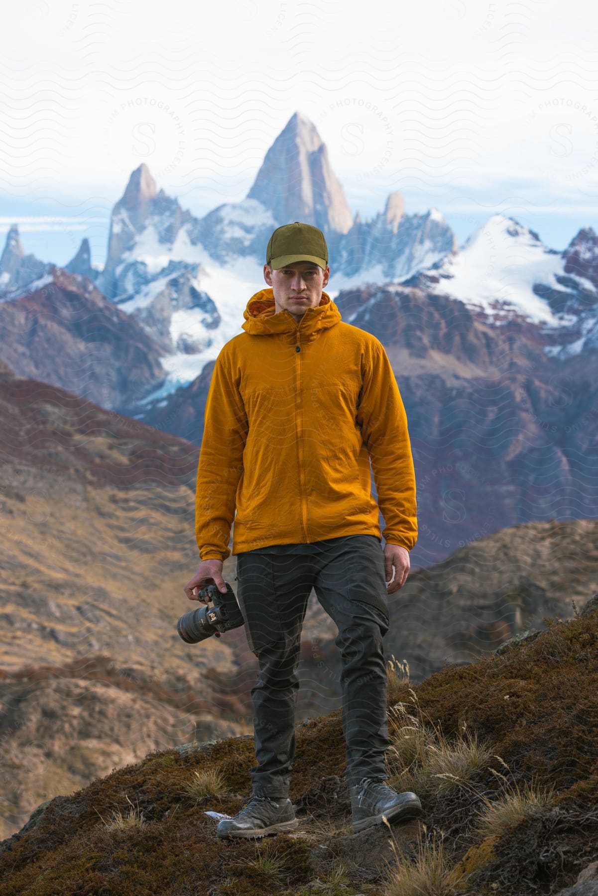 A hiker in an orange windbreaker stands on a grassy hill holding a camera with a snowy mountain range in the distance