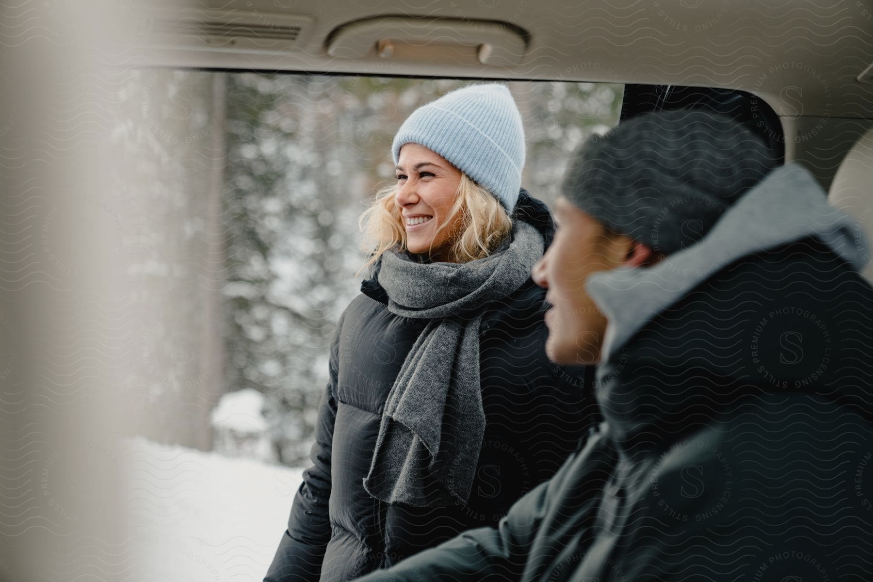 A man and a woman, him inside a car, and her standing outside, smile as he waits for her to get in and take shelter from the snowy weather