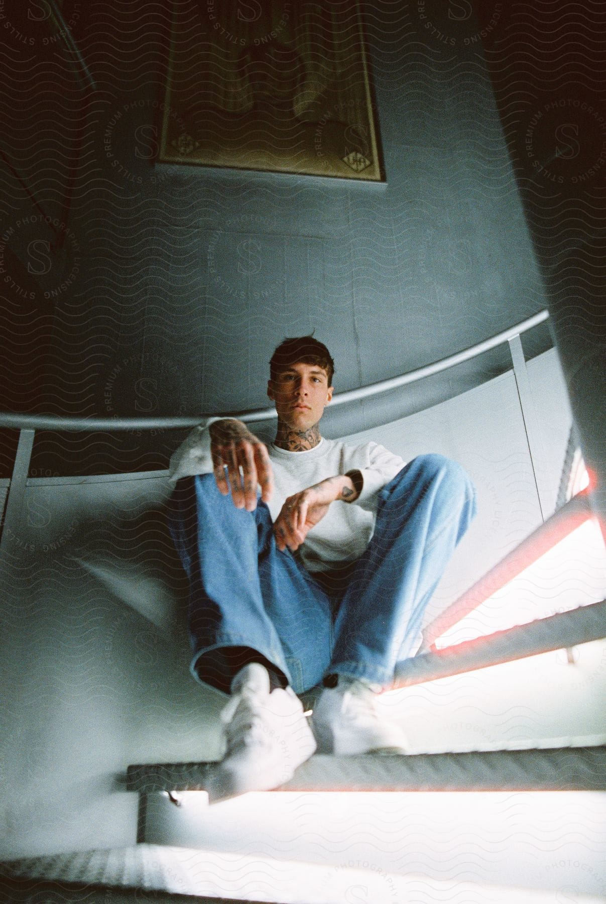 A man with tattoos sits on a stairwell with a painting in the background