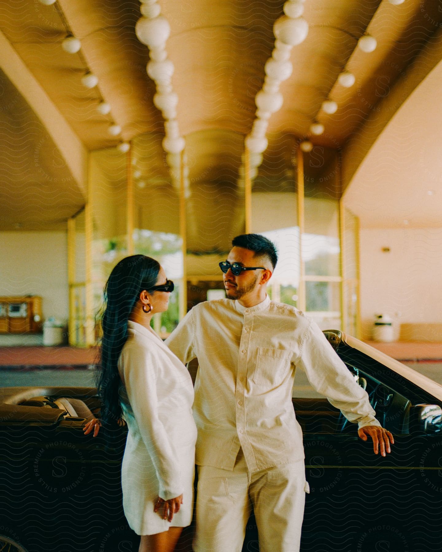 A man and a woman wearing white clothes and sunglasses are looking into each others faces as they stand inside a large room as he leans against furniture