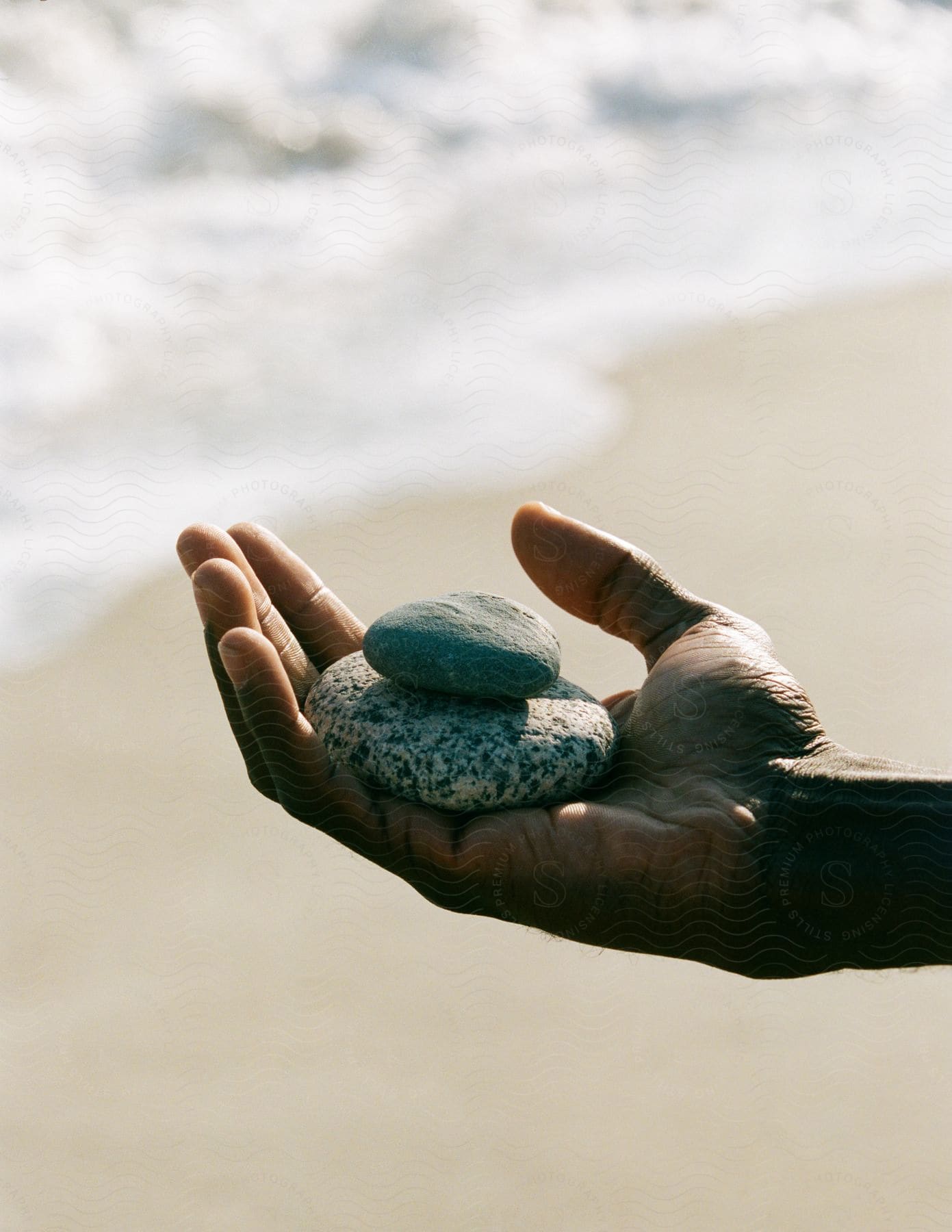 A mans hand holds two stacked oval stones on the beach