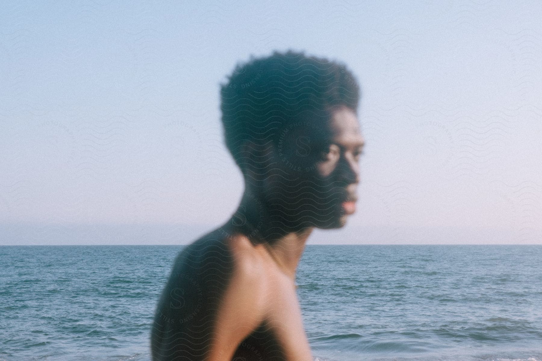 A shirtless black man stands next to the ocean under a blue sky