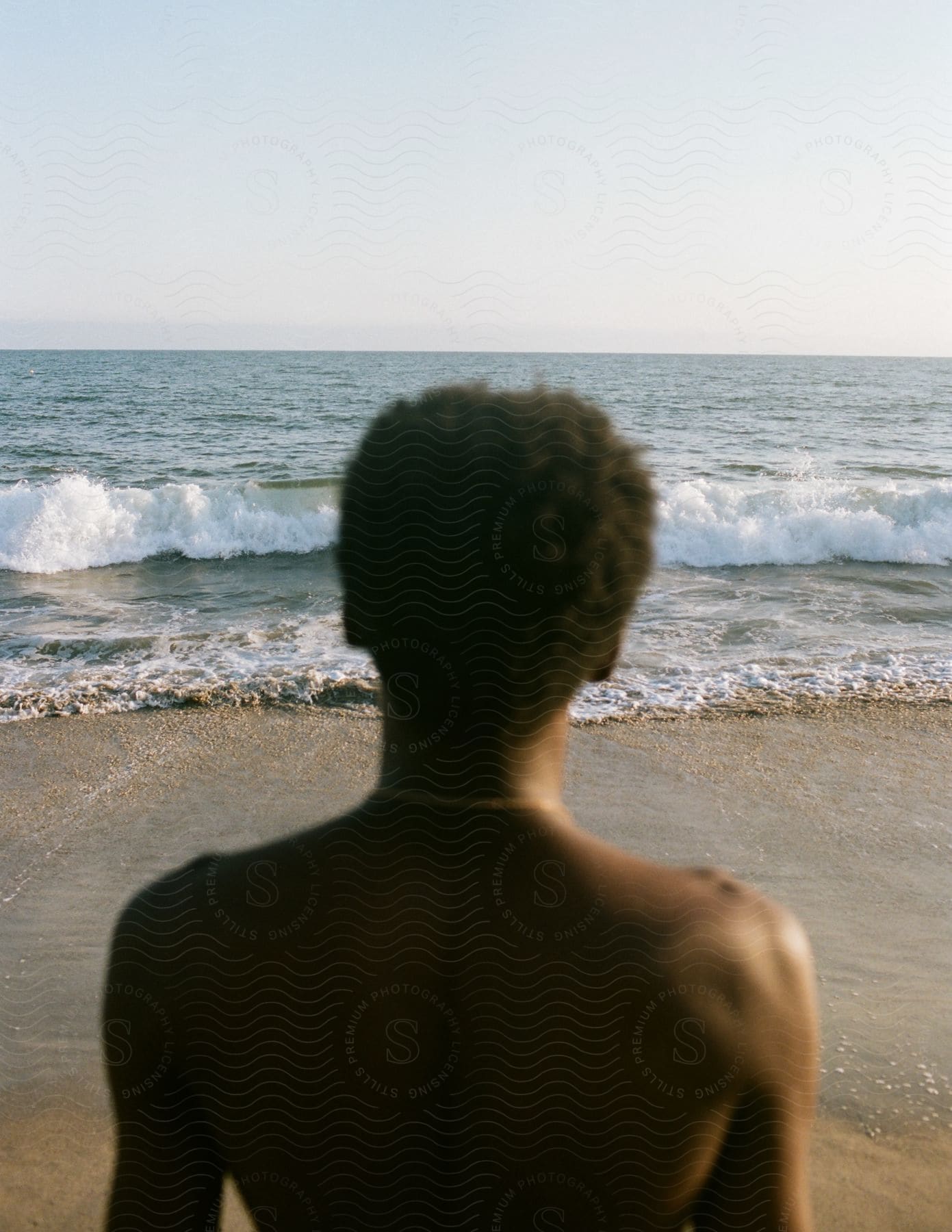 Shirtless young black man standing near the shoreline watching the ocean