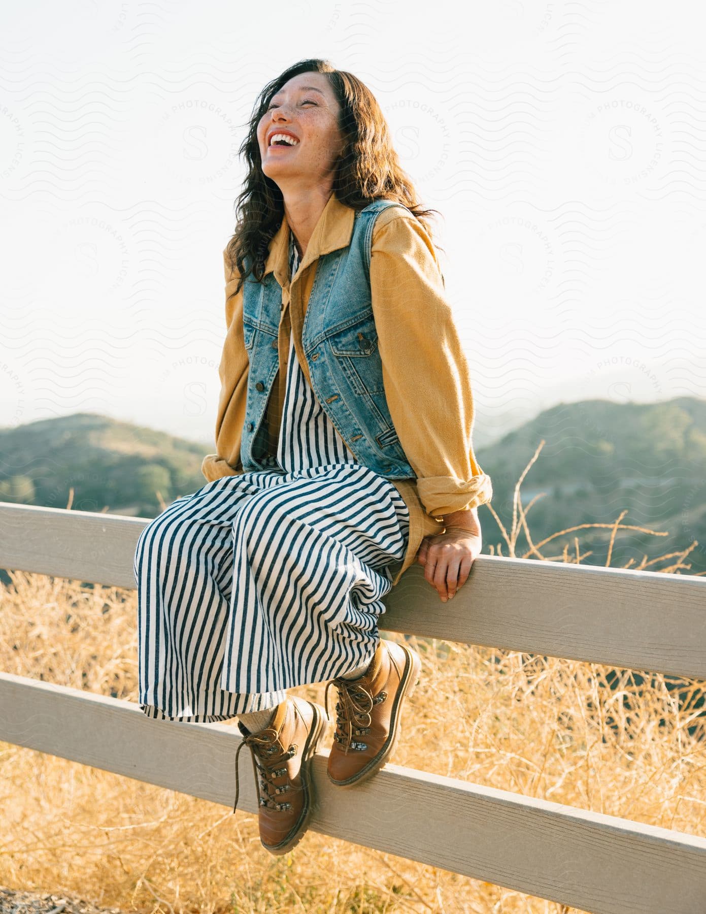 A smiling woman sits on a fence with mountains in the background