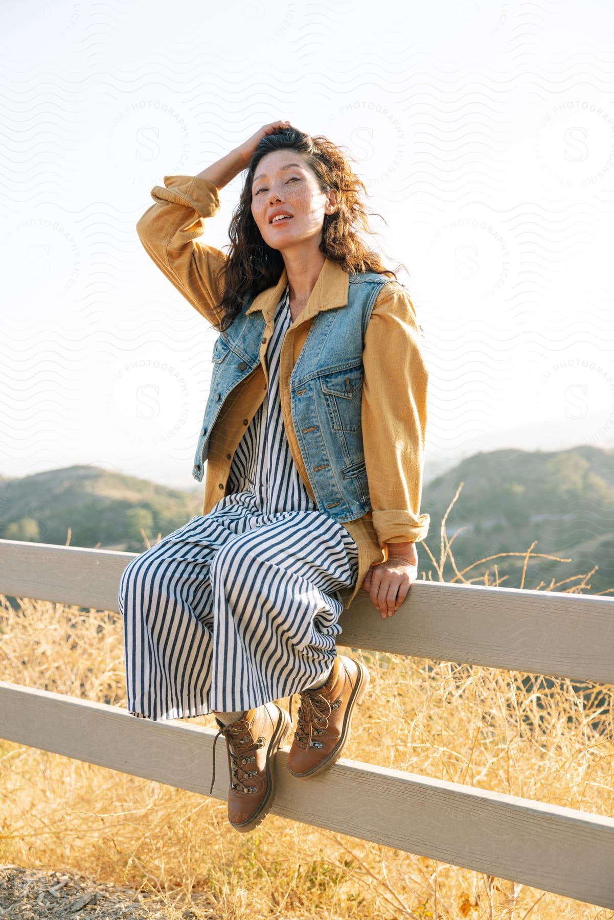 Woman with curly hair sits on wooden fence near farm land touching her hair and smiling in the sunlight