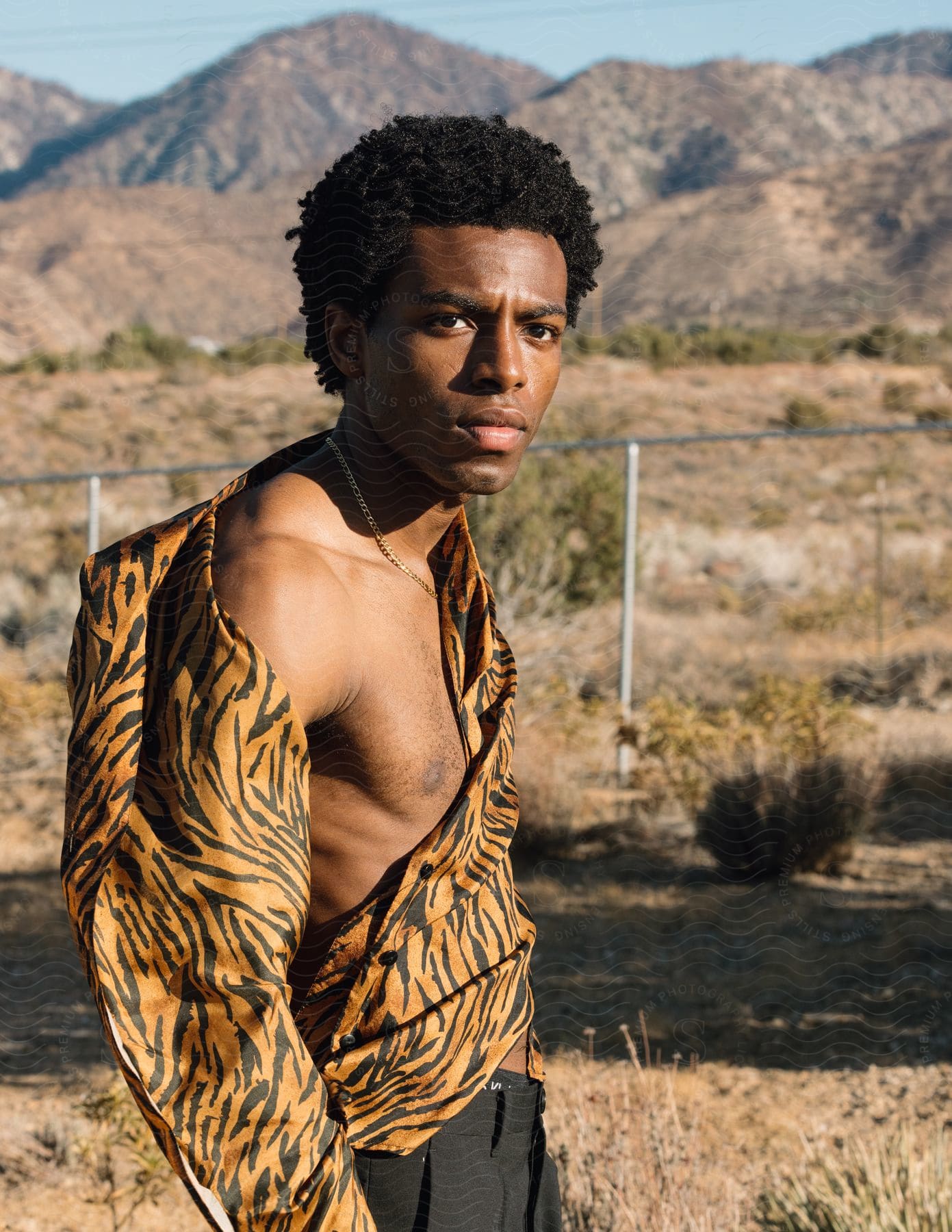 A man with black hair and a tattoo on his skin standing in a desert with plants and a clear sky