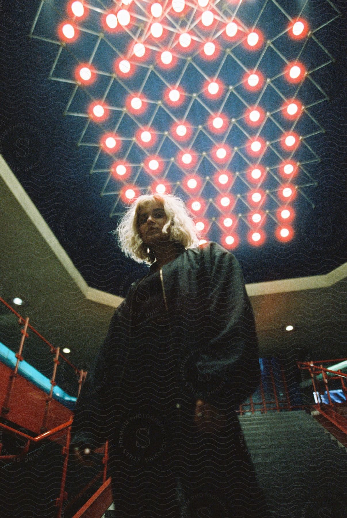 Blond woman in coat stands in front of staircase under red light fixture in building looking down with angry expression