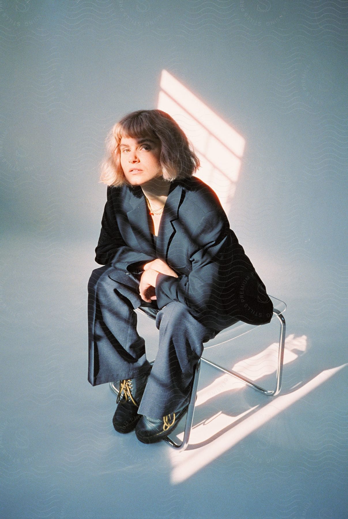 A confident white teenage lady in a stylish suit sits in a chair illuminated by gentle rays of sun passing through a nearby window