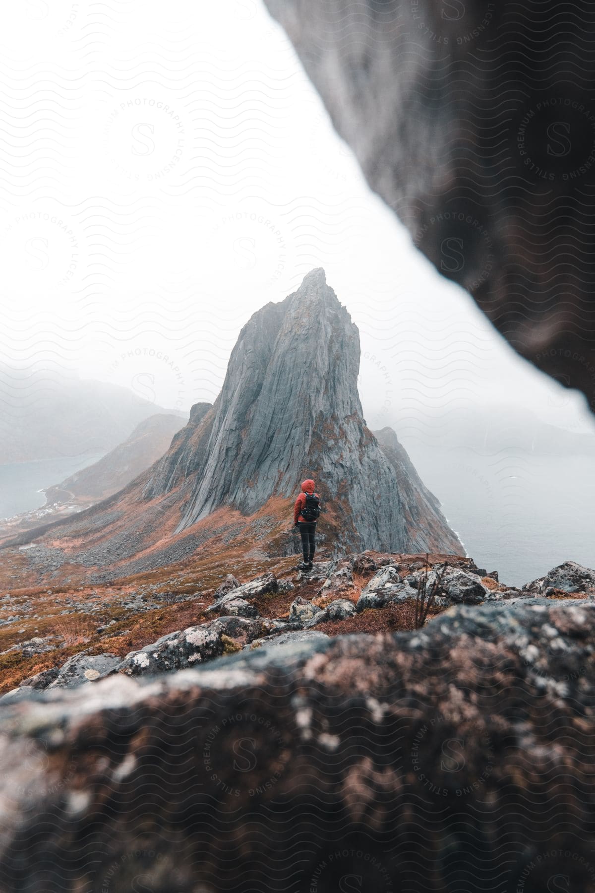 Person standing atop a mountain peak surrounded by fog overlooking a lake