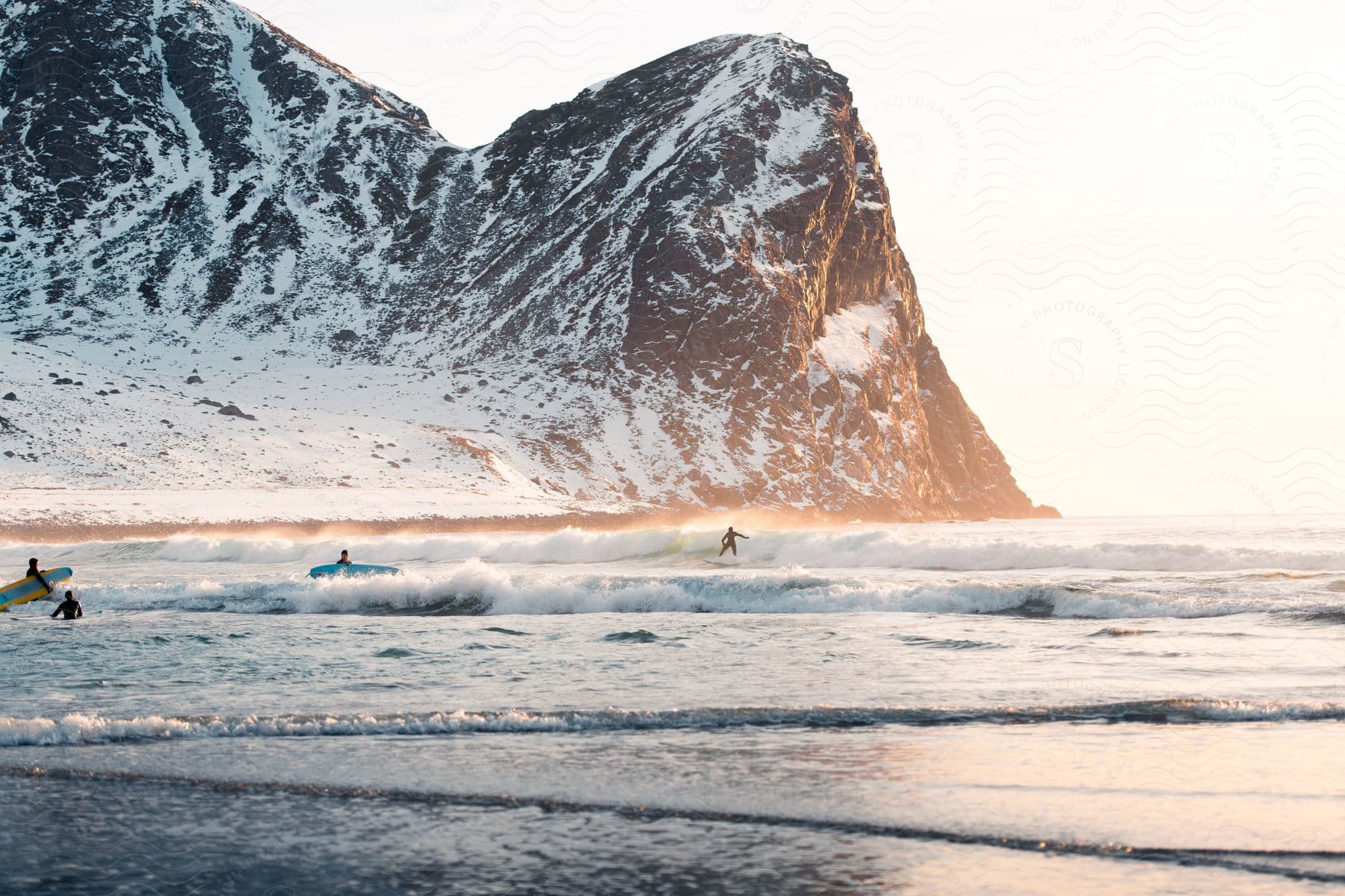 Surfers enjoy waves at a beach with a snowy mountain in the background