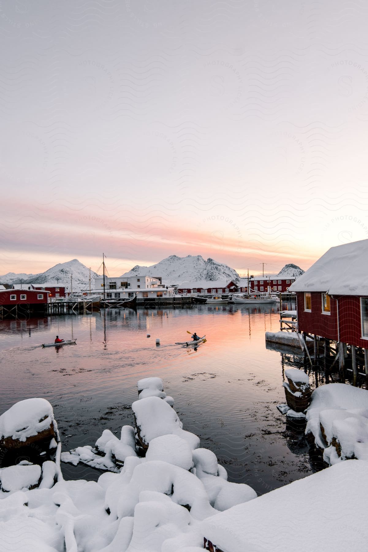 Two men paddling canoes along the water near a coastal city with icy mountains in the background