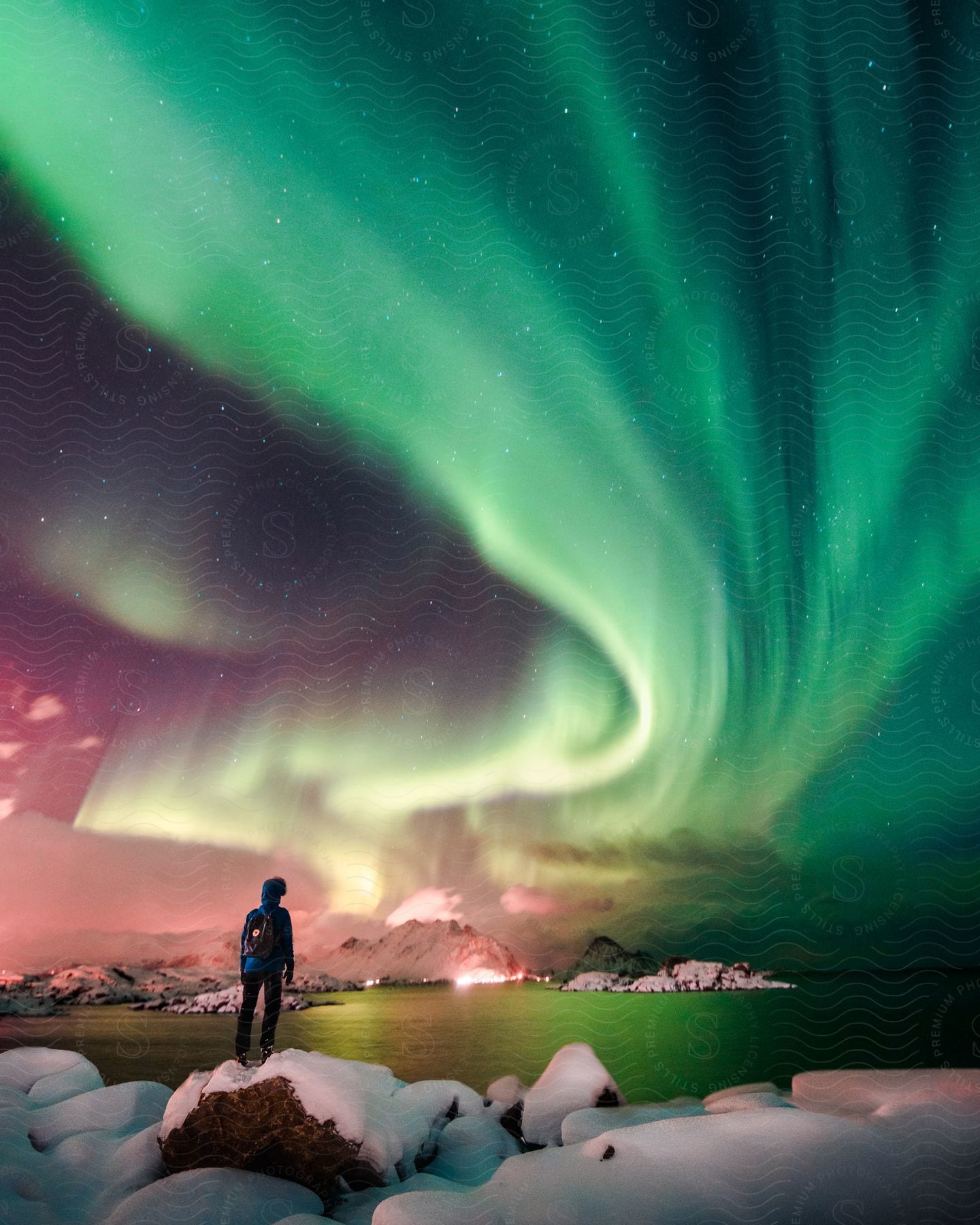 A person stands on a snowy rocky coast in lofoten norway watching the northern lights