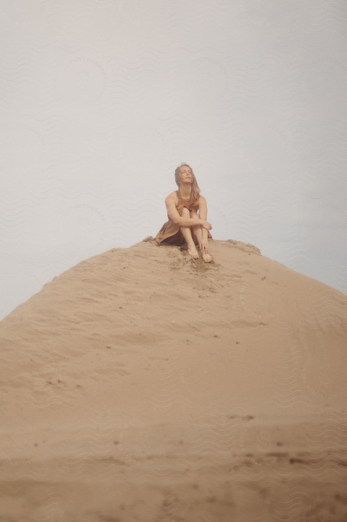 A woman sits barefoot upon a sand dune outdoors during the day