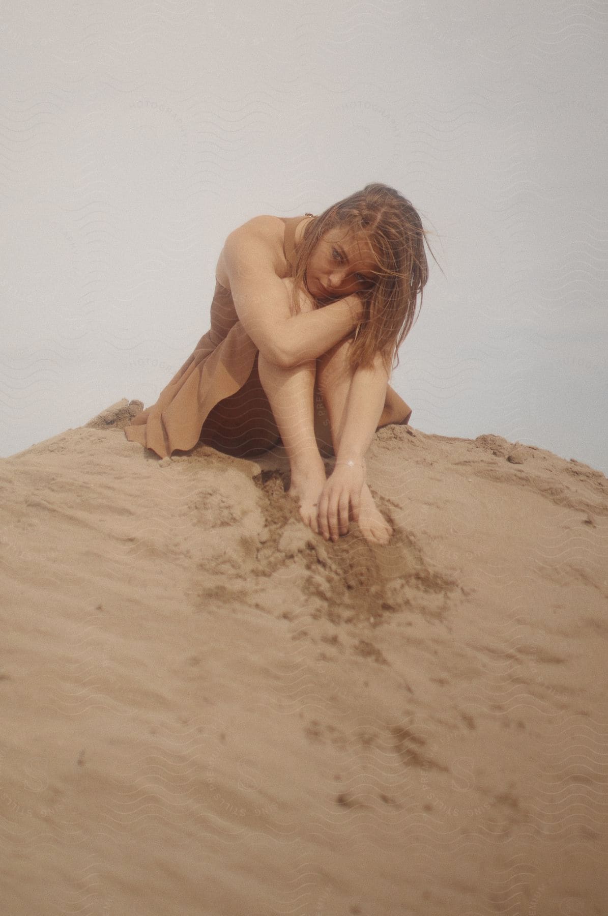 A young woman sits on a sand dune hugging her knees