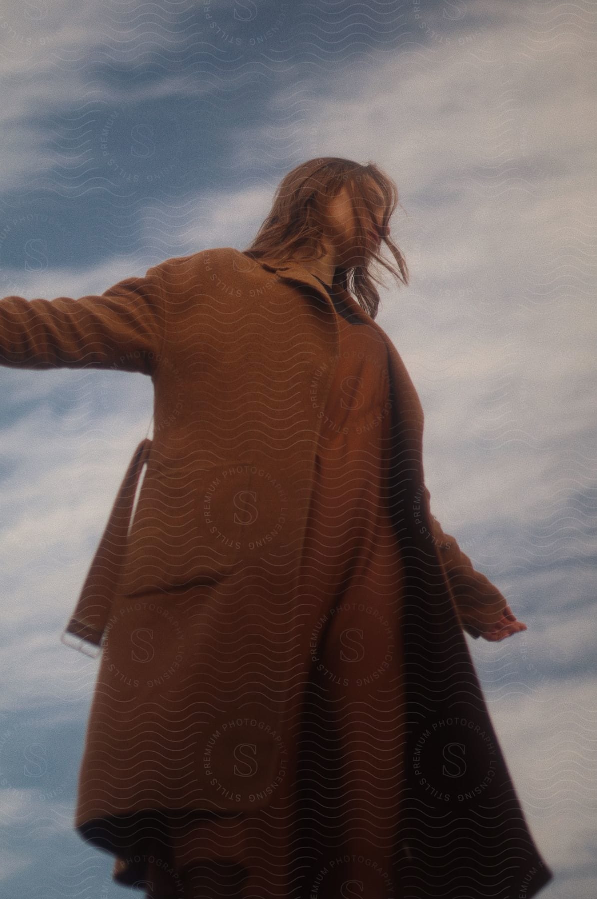 A woman in a brown trench coat and full-body pajamas stands on a cloudy day, her shoulder-length hair blowing in the breeze, arms outstretched to the sky.