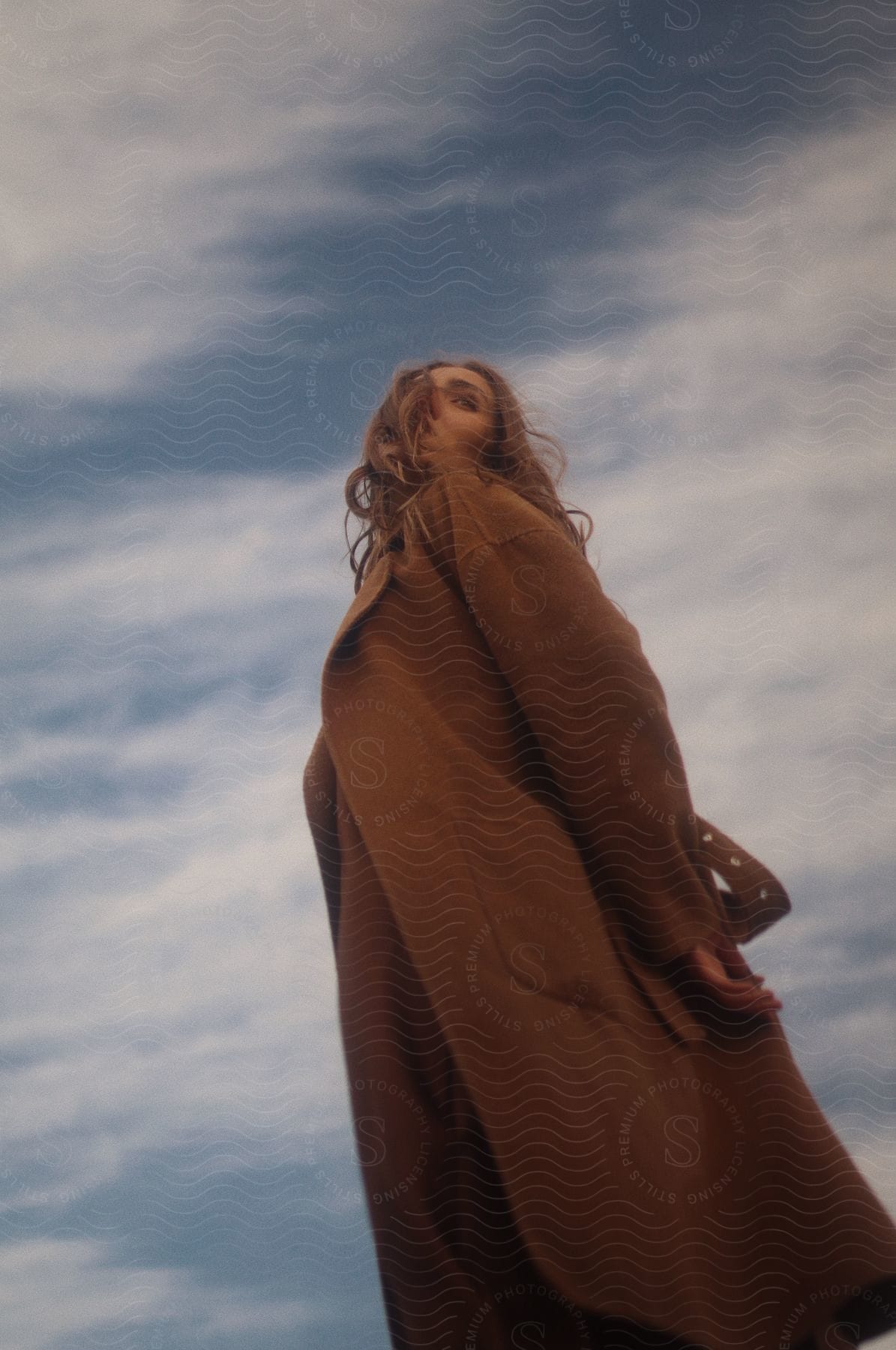 A young woman poses in a long brown coat with cloudy skies in the background.