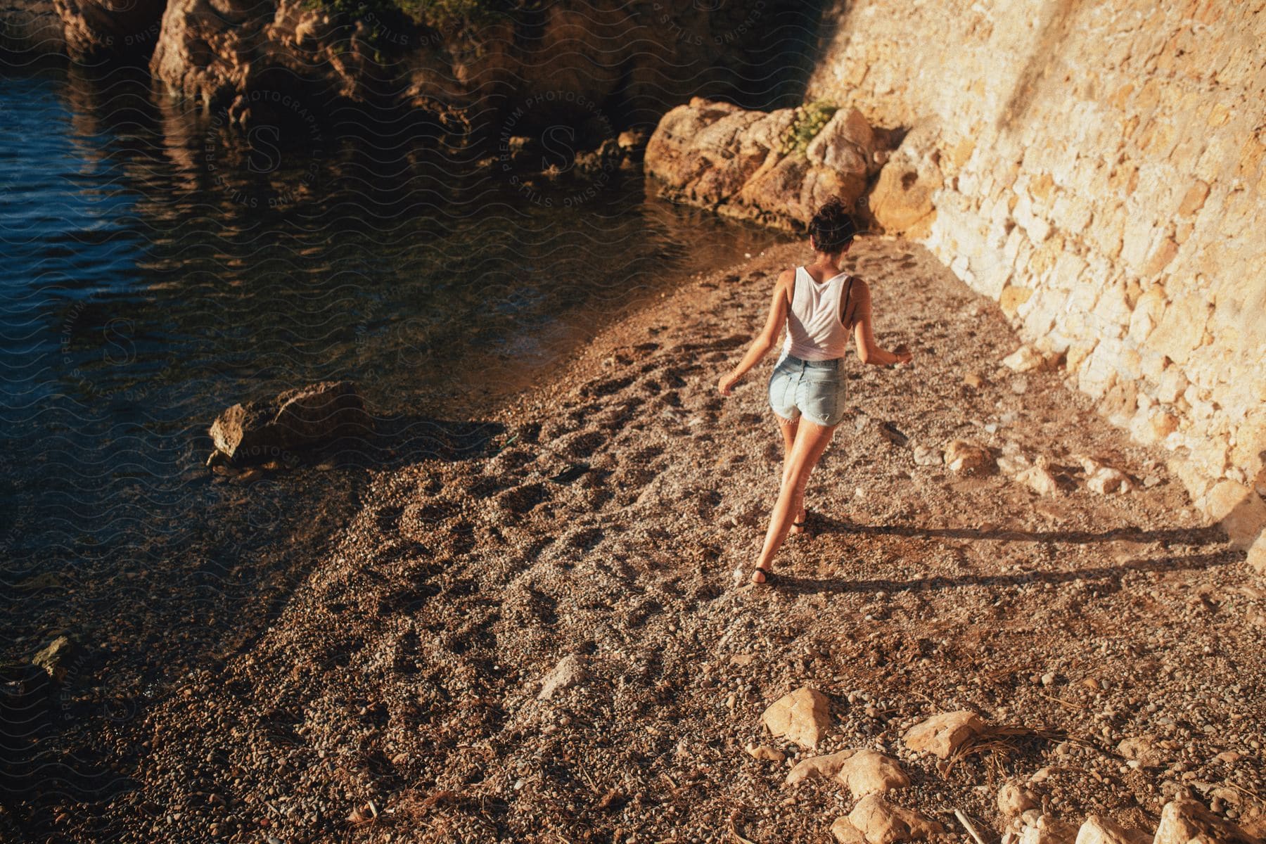 A young woman in a white tank top jean shorts and sandals walks along the beach near a cliff