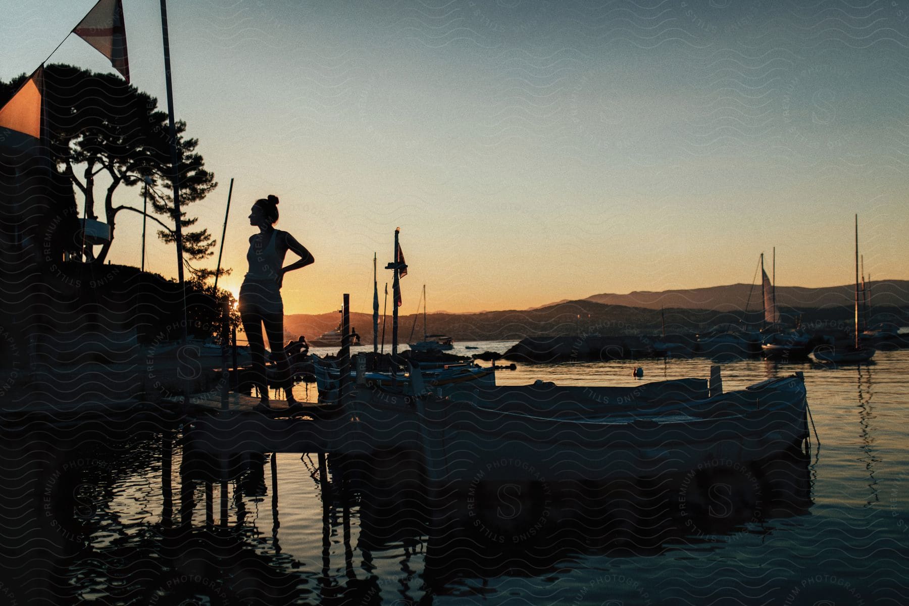 A woman stands on a boat dock near boats in the water as the sun sets