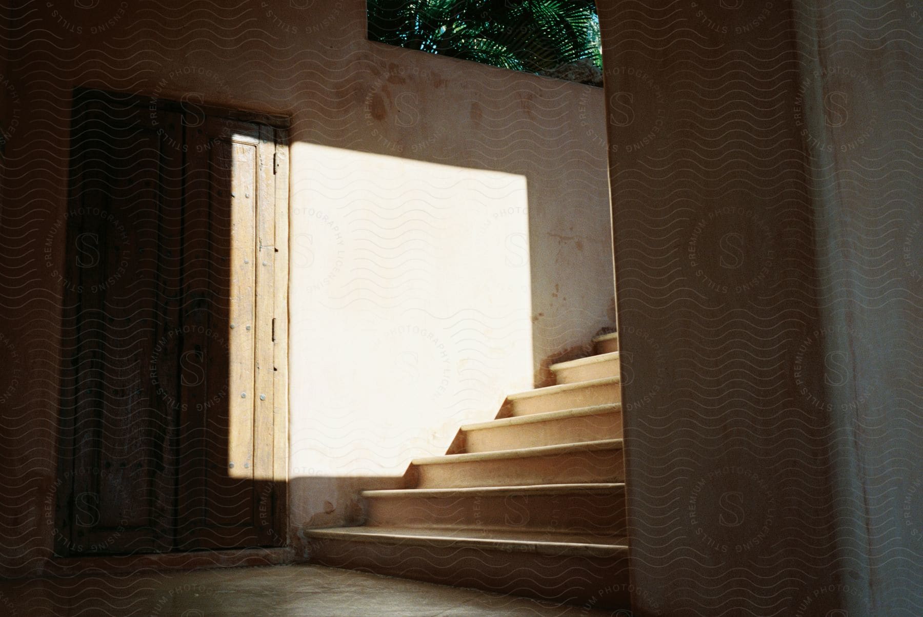 Light shines on a wall and rustic wooden door next to a light wood staircase in mexico