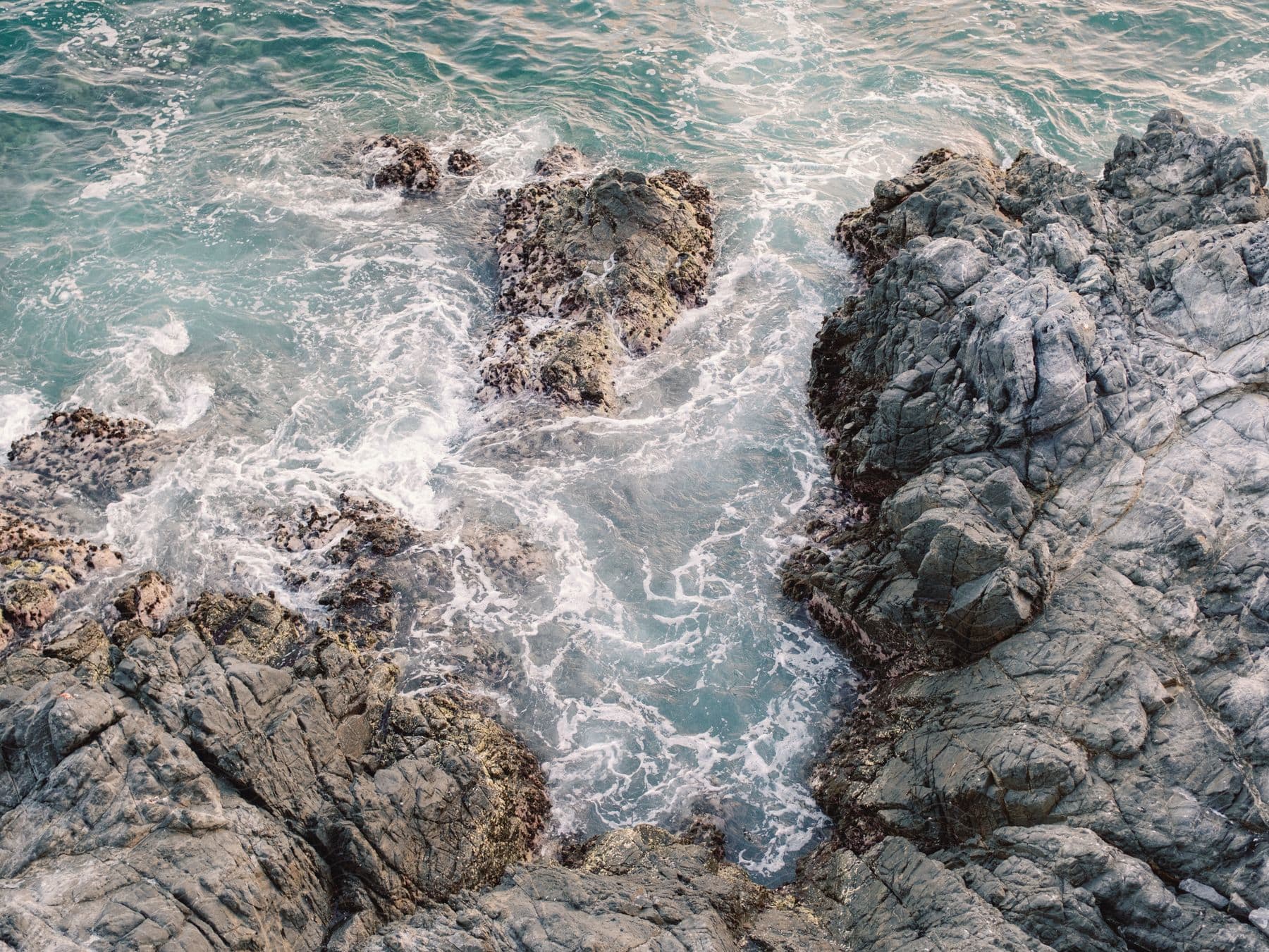 Turquoise waves crash against grey volcanic rocks along the shoreline