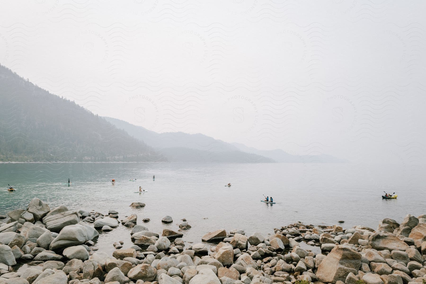 Rocky lake coastline with paddleboarders and kayakers in the distance on a hazy day at lake tahoe