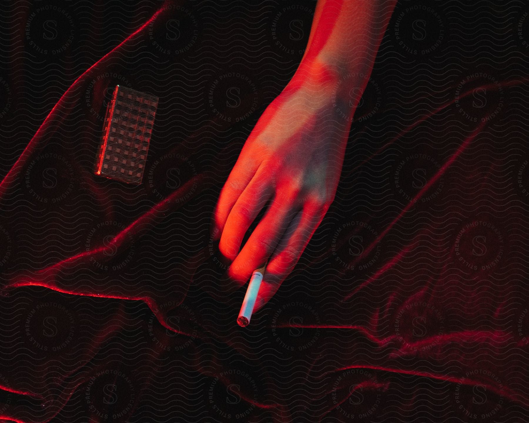A persons arm glows crimson in the reflection of a lamplight holding a cigarette in a bed of deep red velvet