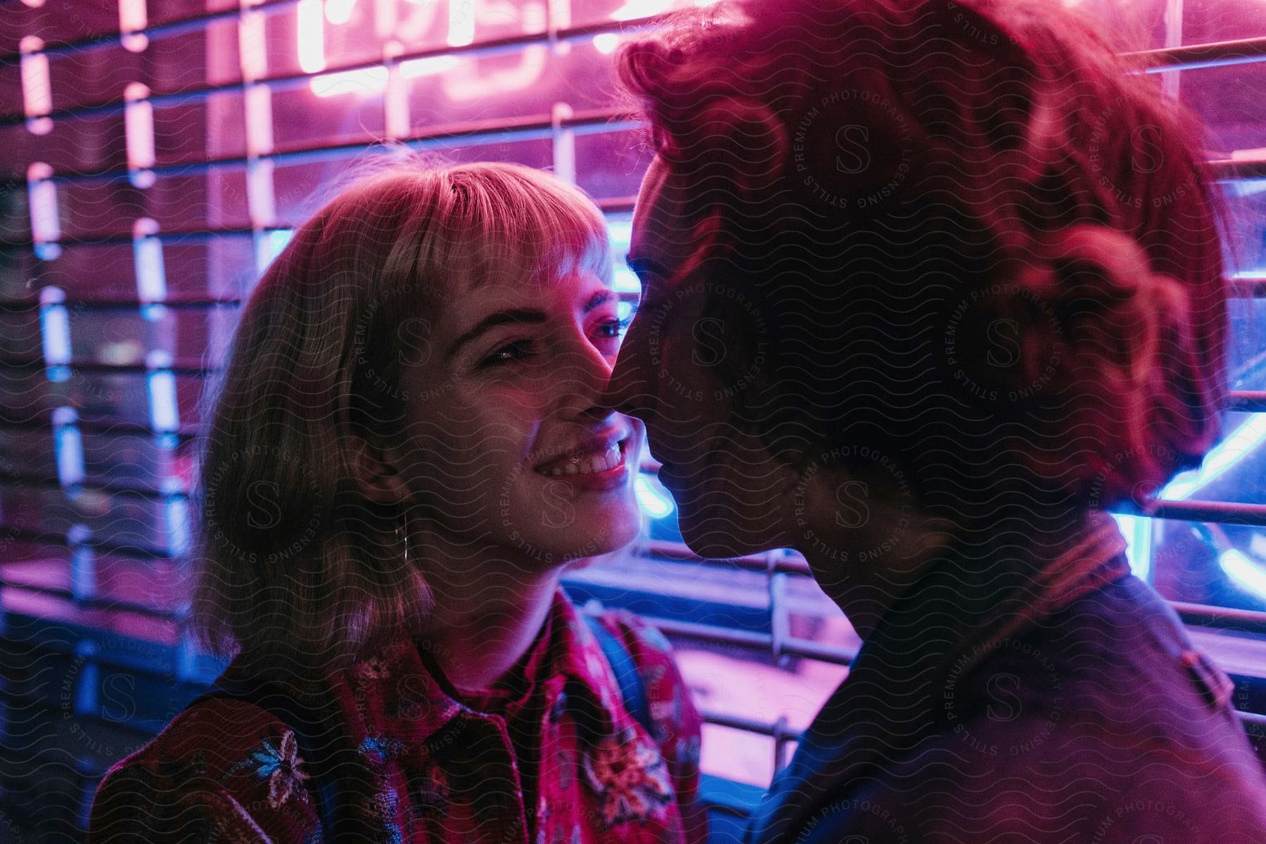 A young couple stand next to a gated storefront with pink and blue florescent light touching noses