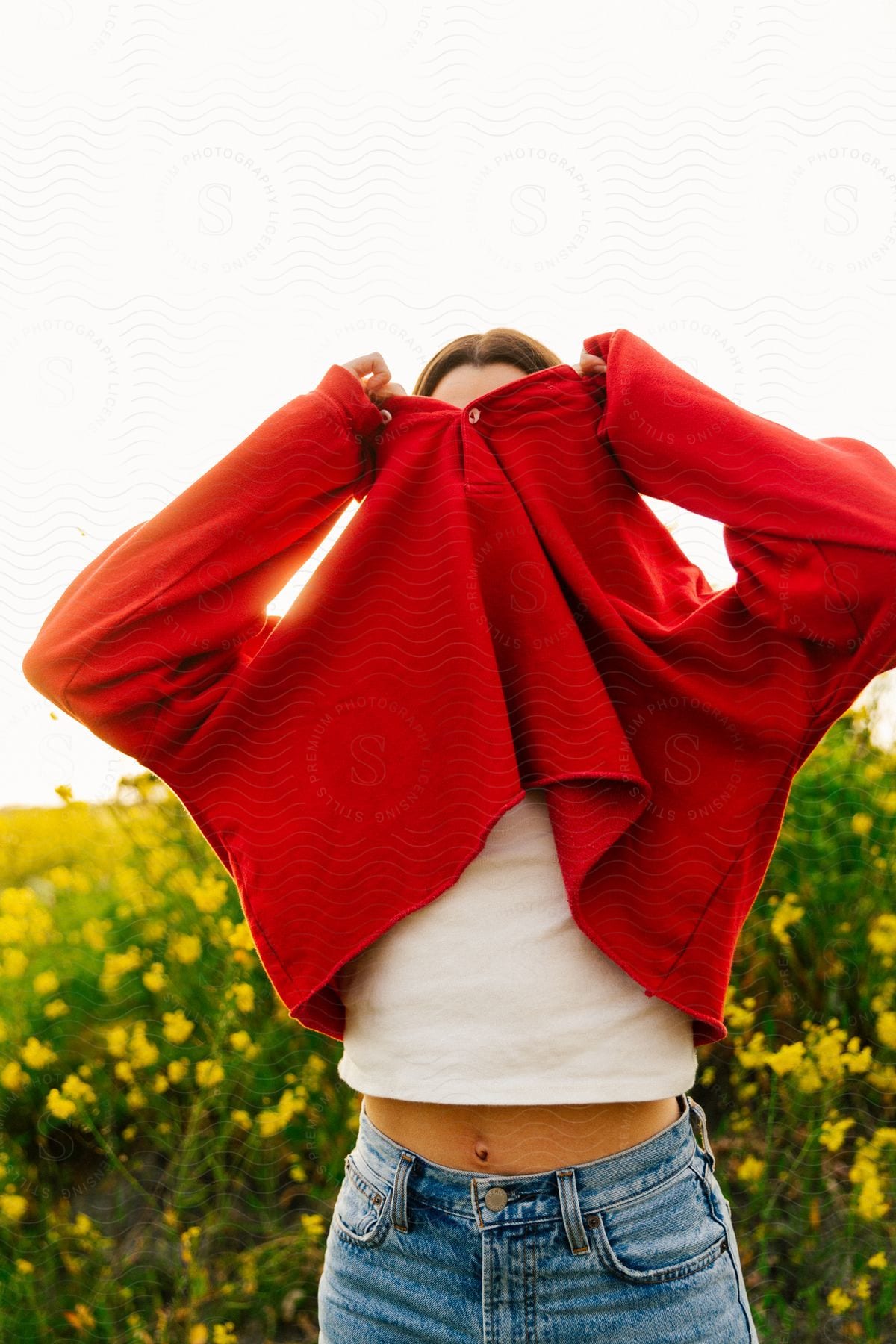 A young woman pulls a red sweatshirt over her head near a field of yellow flowers