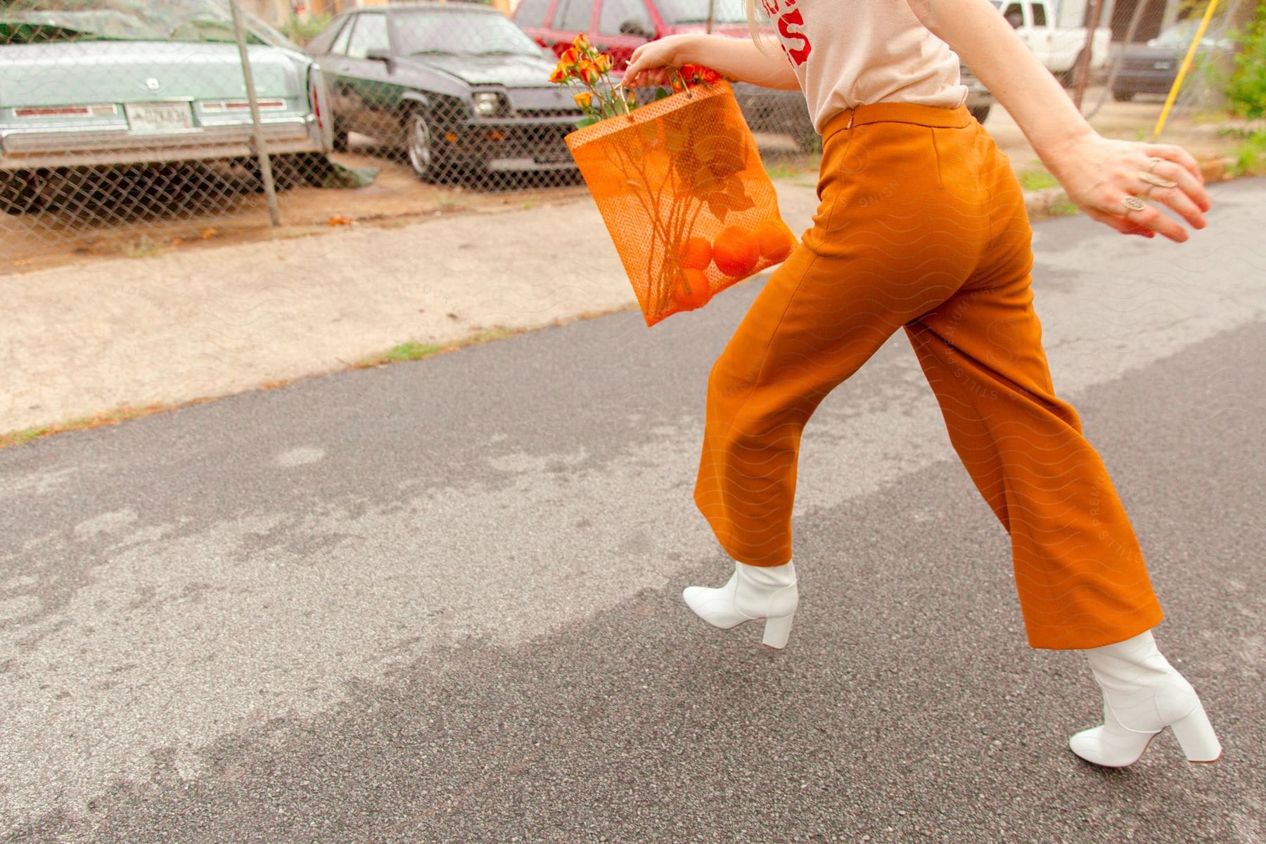 A stylish lady stands on a sunny street wearing vibrant orange trousers holding a fashionable orange bag adorned with flowers and wearing chic white shoes