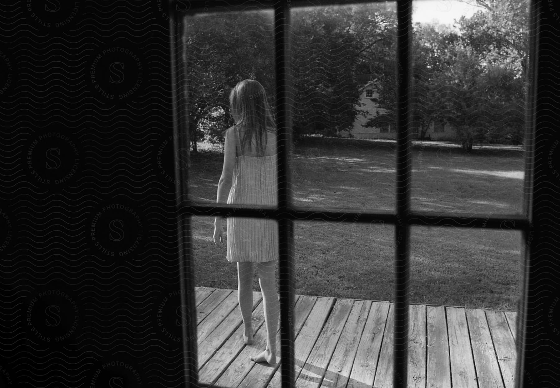 A barefoot girl stands on a deck looking out a window