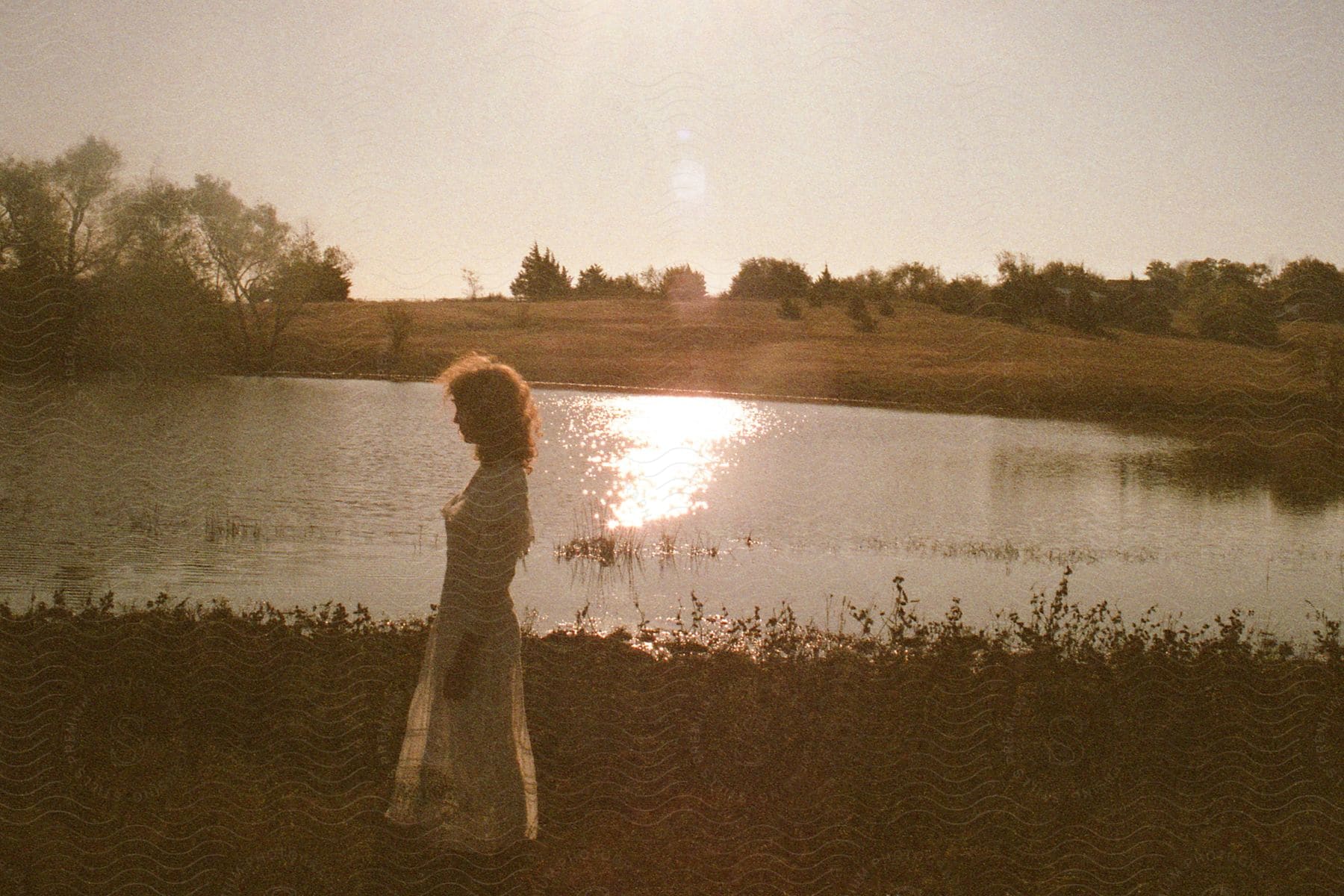 A Woman In A White Gown Walks Along A Riverside Surrounded By Lush Trees And Serene Beauty