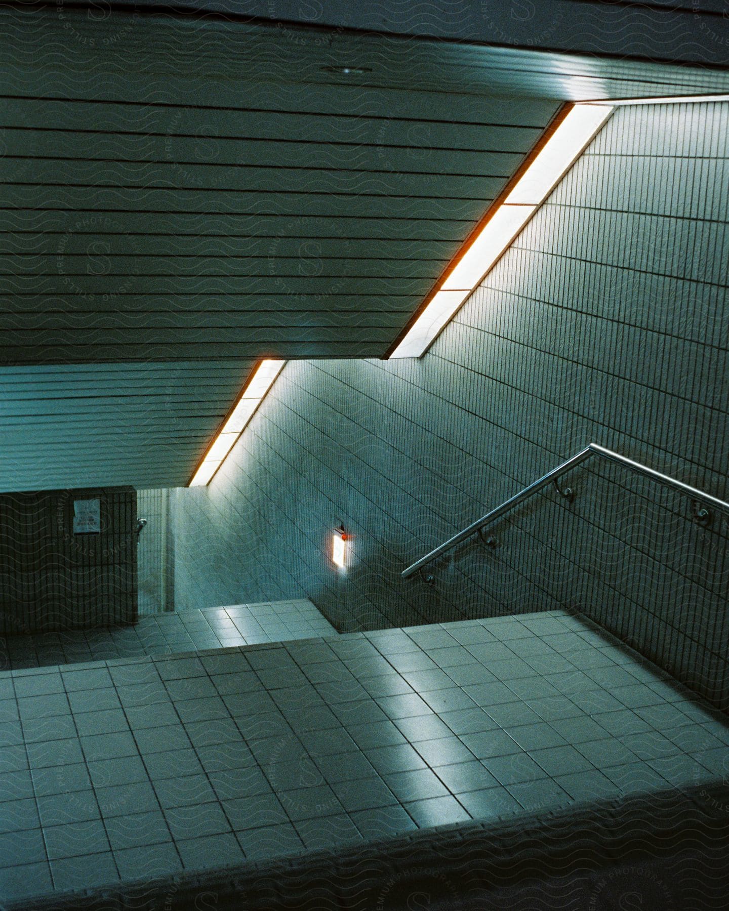 A lit stairway with silver handrails and tile on the landing floor with a partially open gate