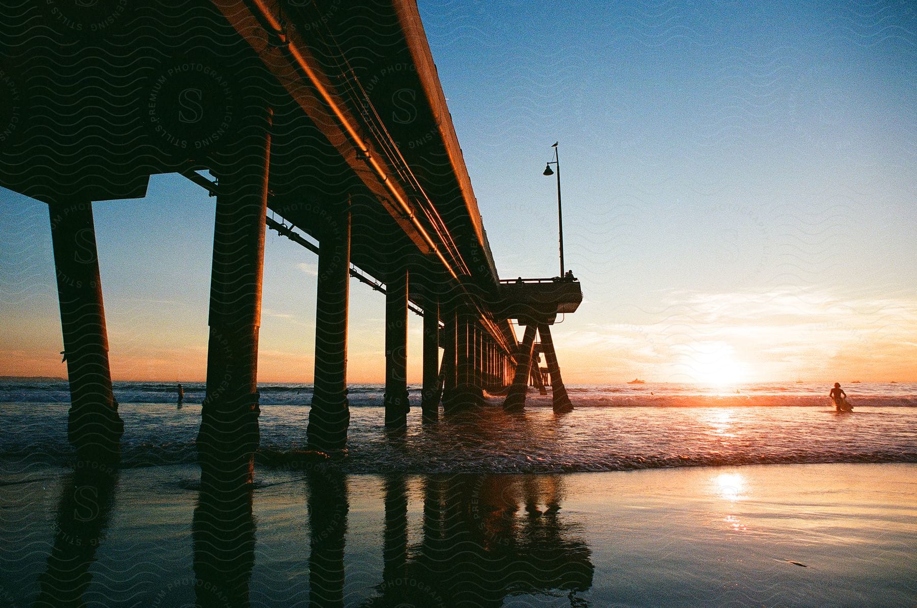 A wooden pier on a beach at sunset with two people coming out of the water