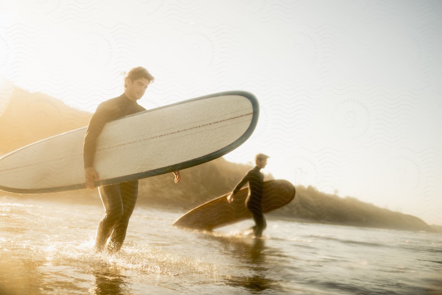Two surfers in wetsuits walk in ankle deep ocean water while carrying their surfboards.