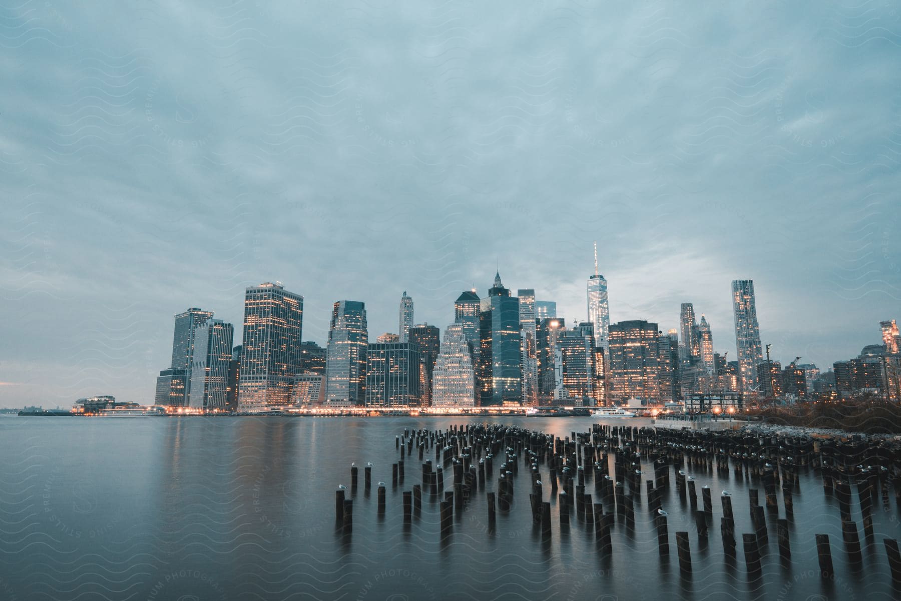 A lake with wooden stakes in front of a lit city with towers and buildings