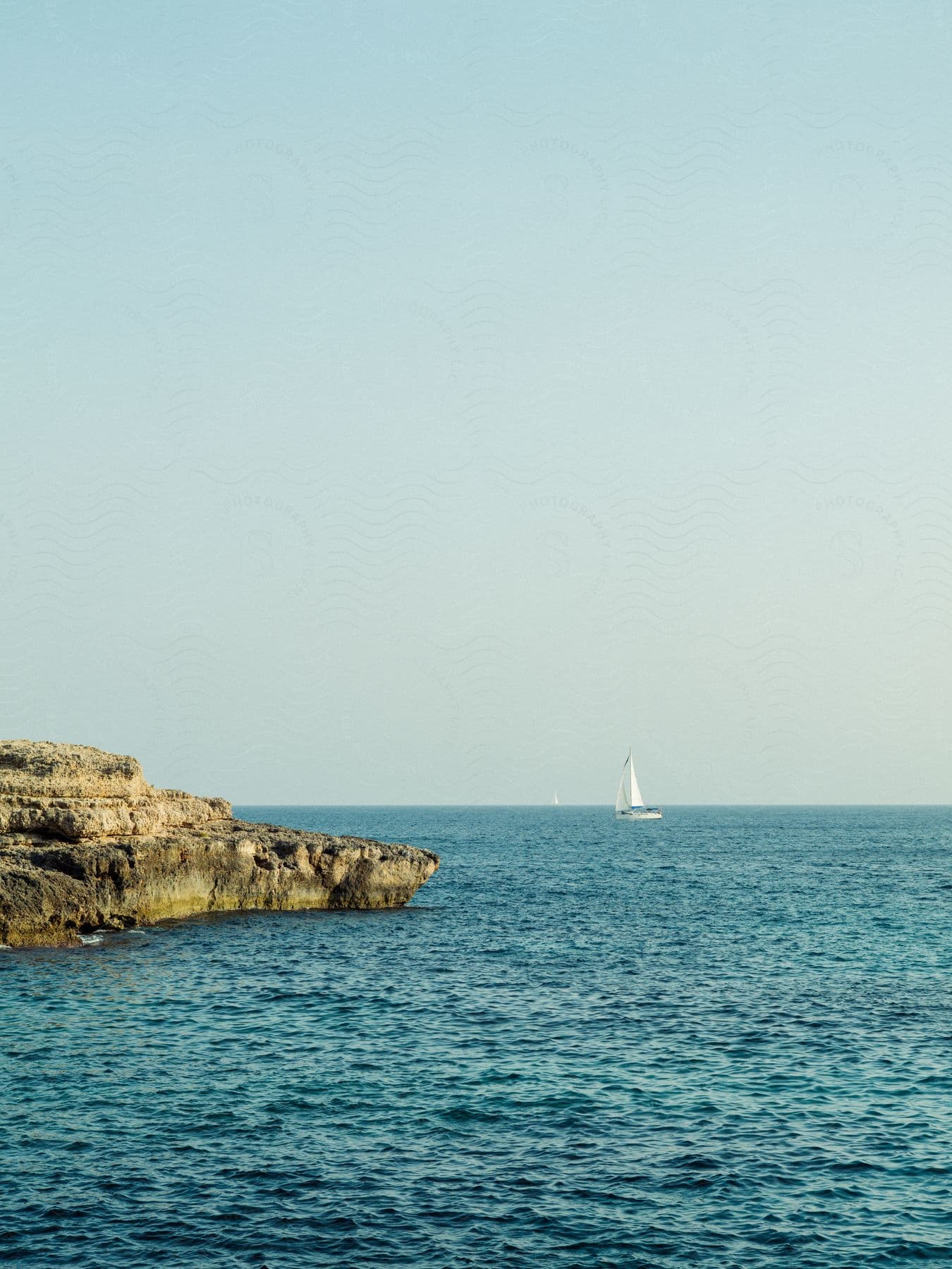 Sailboats float across the water off a rocky coast under a clear sunny sky